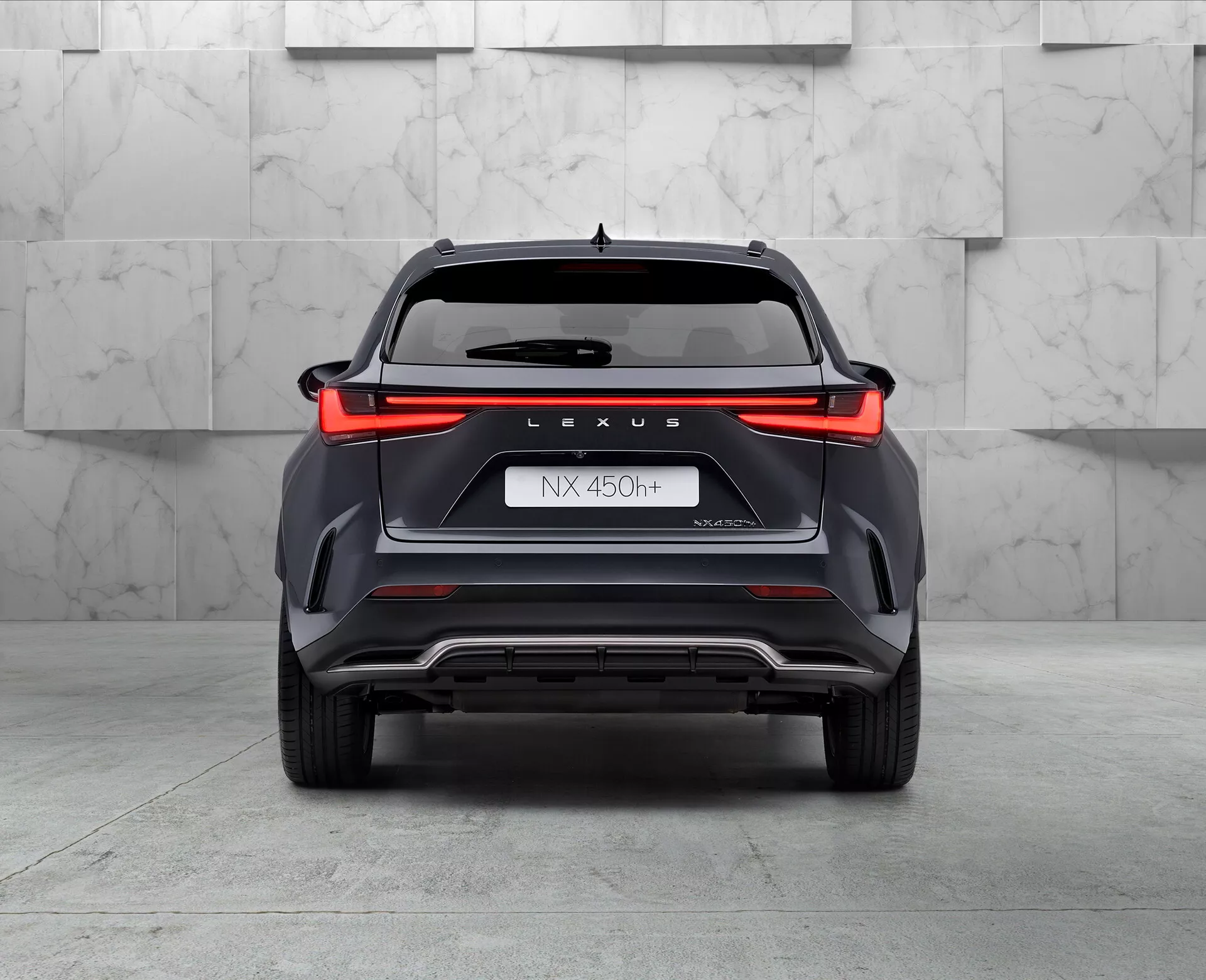 Pohled zezadu na hybridní SUV Lexus NX 450h, snímek pořízený z nízkého úhlu lehce nakloněného doleva, zobrazující plnší šíři zadního světla, značku 'LEXUS', označení modelu 'NX 450h', název 'NEXUS' a symbol hybridu 'h'. Nárazník s úhlovými stylingovými prvky, ventilačními elementy a stříbrnou kluznou deskou, viditelný levý výfukový konec. Pozadí s leštěnými podlahami a neutrálne zbarvenou stěnou, evokující prostředí showroomu.