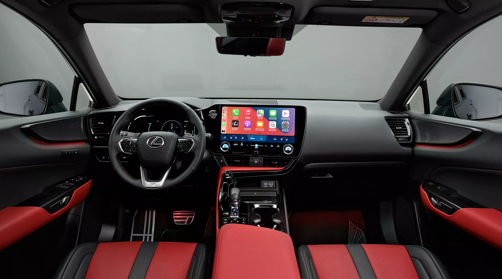 Interiér Lexus NX 2021 zaměřený na přední kabinu, pořízený z centrální pozice. Širokoúhlý pohled ukazuje palubní desku s velkou centrální dotykovou obrazovkou infotainmentu zobrazující mobilní rozhraní, fyzické ovládací prvky klimatizace a řadicí páku automatické převodovky. Multifunkční volant s ovládacími tlačítky naznačuje pokročilé funkce. Sedadla jsou čalouněná dvoubarevnou červenou a černou kůží, zajišťující sportovní vzhled. Dveřní panely odpovídají červeno-černému motivu s ovládacími prvky elektrických oken a viditelnou mřížkou reproduktoru, zdůrazňující modernitu a luxus.