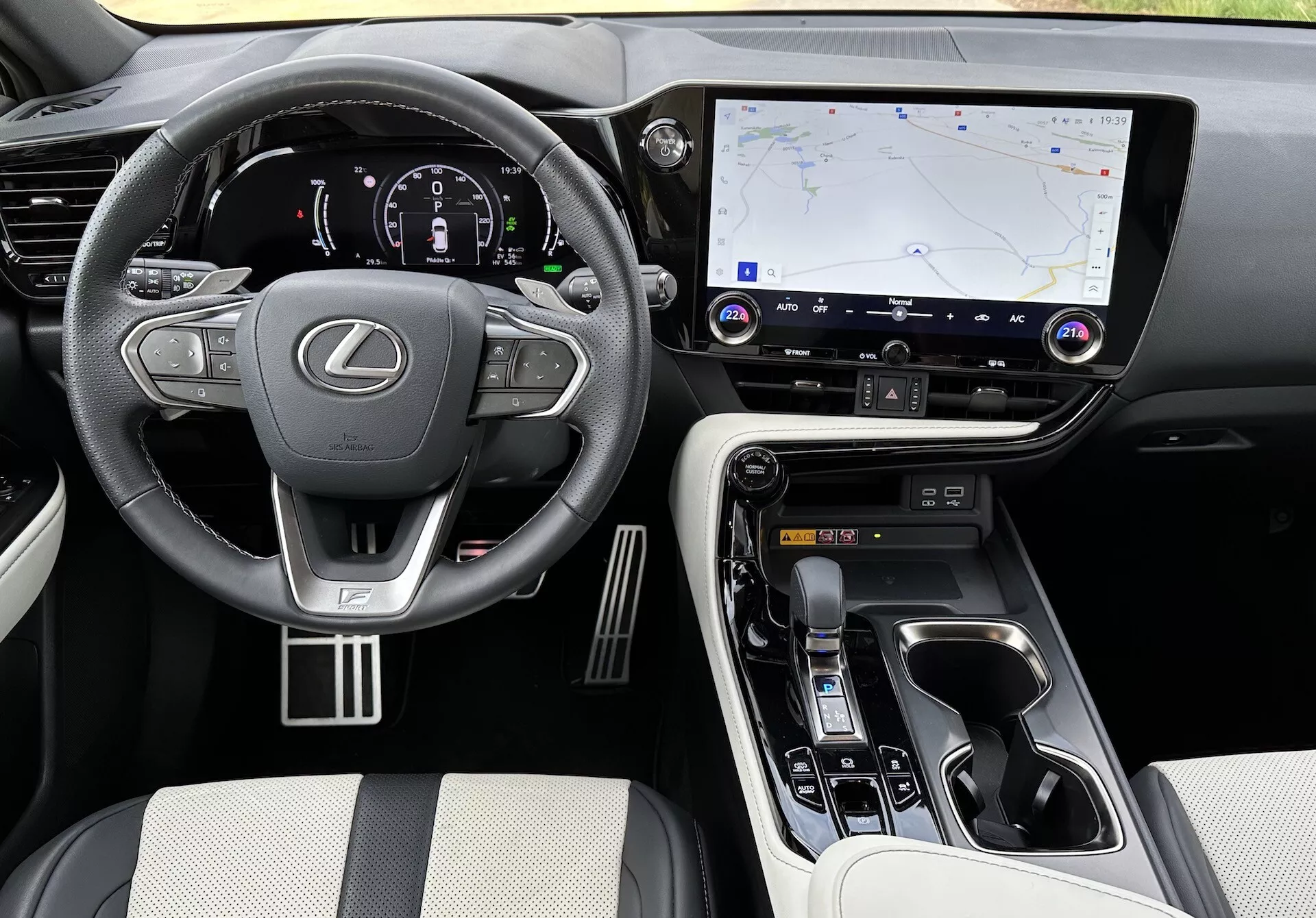 Interiérový pohled na Lexus NX 2021 zvýrazňující oblast řidiče a středovou konzolu zvýšeného úhlu, s logem Lexus na volantu s ovládacími prvky, osvětleným digitálním přístrojovým panelem, velkým dotykovým infotainment systémem s GPS navigací, tlačítky pro ovládání klimatizace a zvuku, ergonomickým selektorem převodů, ovládacími prvky režimu jízdy, luxusními materiály na sedadlech a dotykových bodech, kovovými akcenty a viditelnými držáky na nápoje a úložným prostorem.