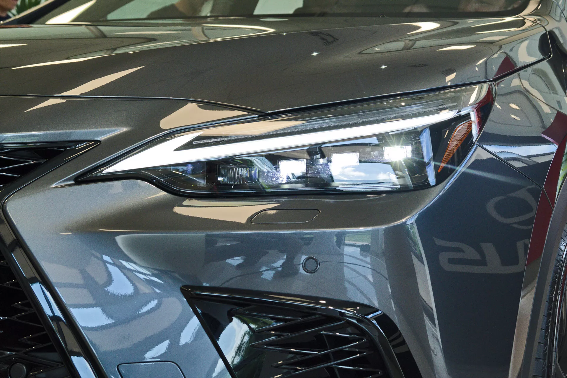 Detailní pohled na přední část modelu Lexus NX 2021, zaměřující se na světlomet na straně řidiče s pokročilou LED osvětlovací technologií a část agresivně navržené mřížky. Obrázek zachycuje odrazivý lesk kovového stříbrného laku a dynamické linie přispívající k aerodynamické estetice vozidla. Fotografováno v interiéru, pravděpodobně v showroomu, zdůrazňující luxusní design a vysoce kvalitní povrchovou úpravu vozidla.