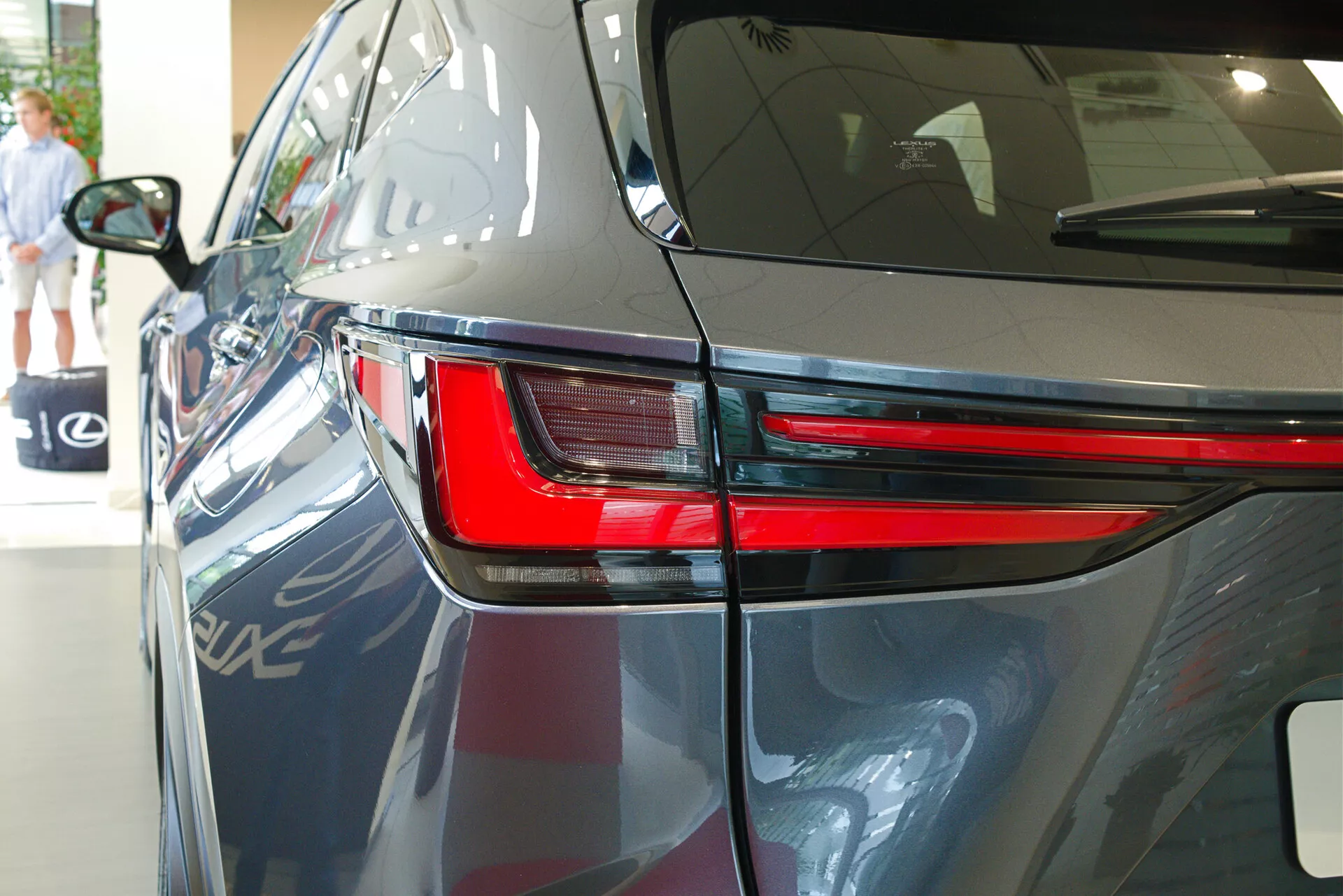 Blízký pohled na Lexus NX 2021 zezadu ve tři čtvrtiny úhlu, který zdůrazňuje elegantní design LED zadního světla, část zadního čtvrtpanelu a část víka kufru. Obrázek ukazuje lesklý tmavý lak vozu, aerodynamické linie karoserie a naznačuje vnitřní výstavní prostor s odrazy dalšího vozidla, architektonickými prvky a osobou kráčející v pozadí.