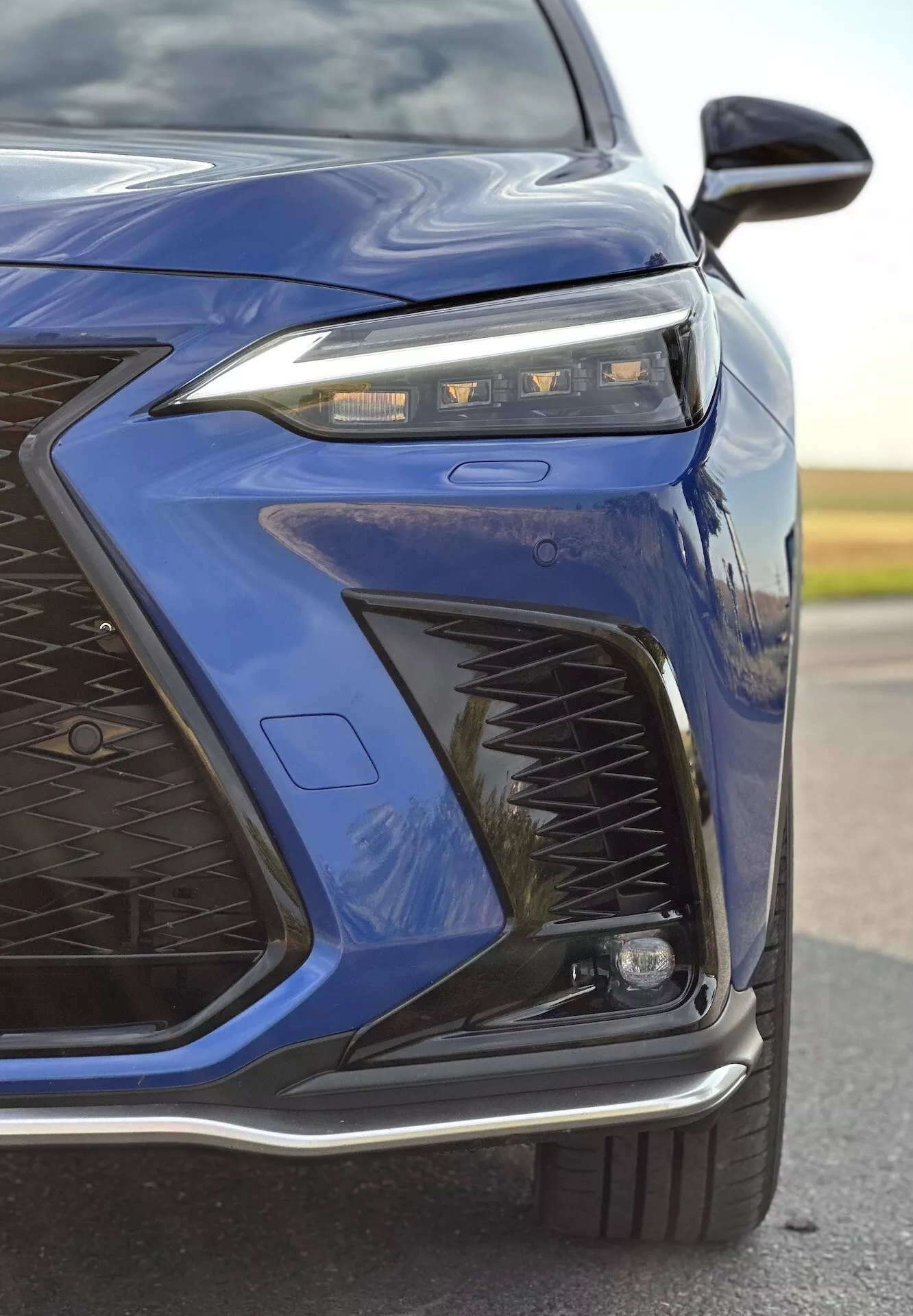 Detailní pohled na přední levý roh tmavě modrého Lexusu NX z roku 2021 s důrazem na pokročilý design LED světlometů, složitý vzor mřížky, sportovně laděný přední nárazník a možné adaptivní osvětlení a aerodynamické prvky s lesklým kovovým povrchem