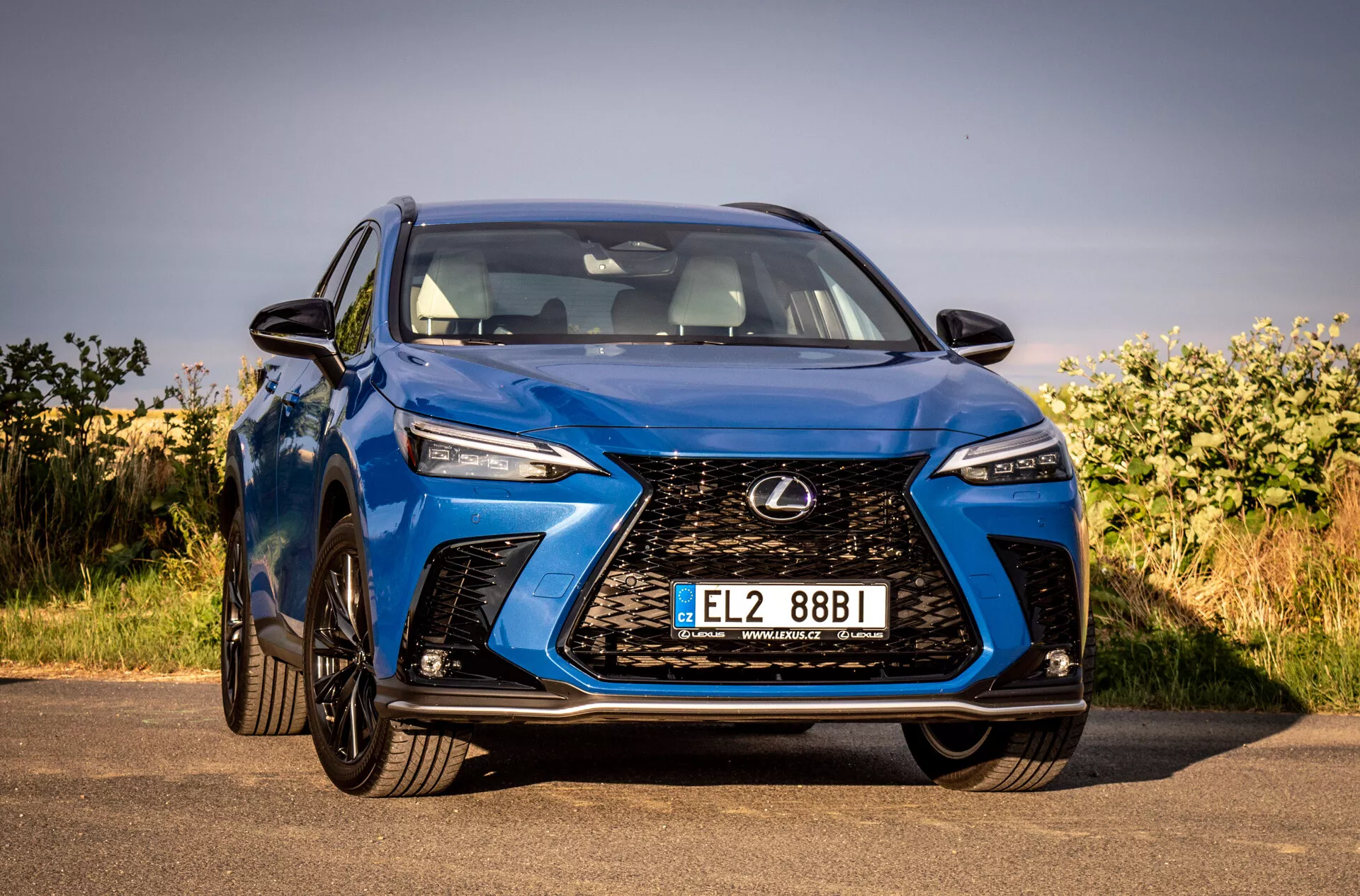 Pohled z předního úhlu na modré SUV Lexus NX 2021 zdůrazňující jeho složitý vřetenový mříž, úhlové LED světlomety, lesklý povrch a aerodynamický design, zaparkovaný venku pod jasnou oblohou