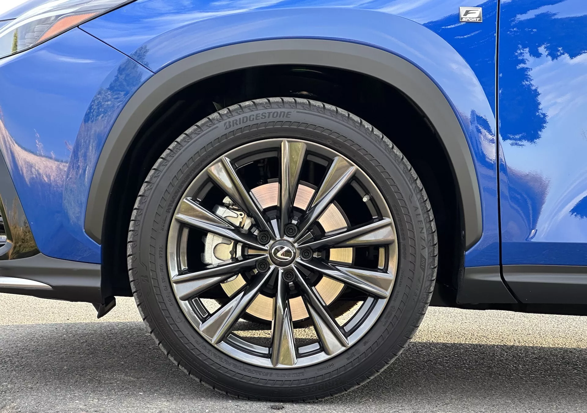 Detailní pohled na přední kolo na straně spolujezdce Lexus NX 2021 s vícepaprskovým designem s dvoubarevným povrchem a pneumatikou Bridgestone, na pozadí s výrazně modrým lakem