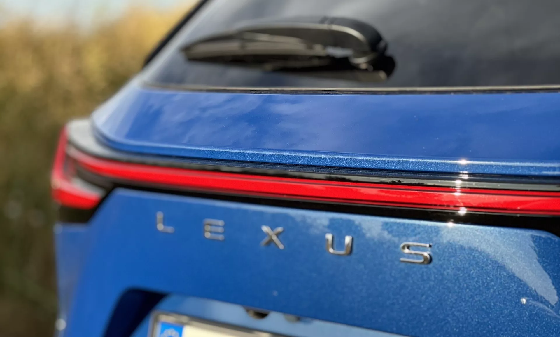 Detailní pohled na zadní část značení vozu Lexus NX 2021 s stříbrným odznakem Lexus, červeným světelným pruhem nad názvem na kovově modrém těle za denního světla