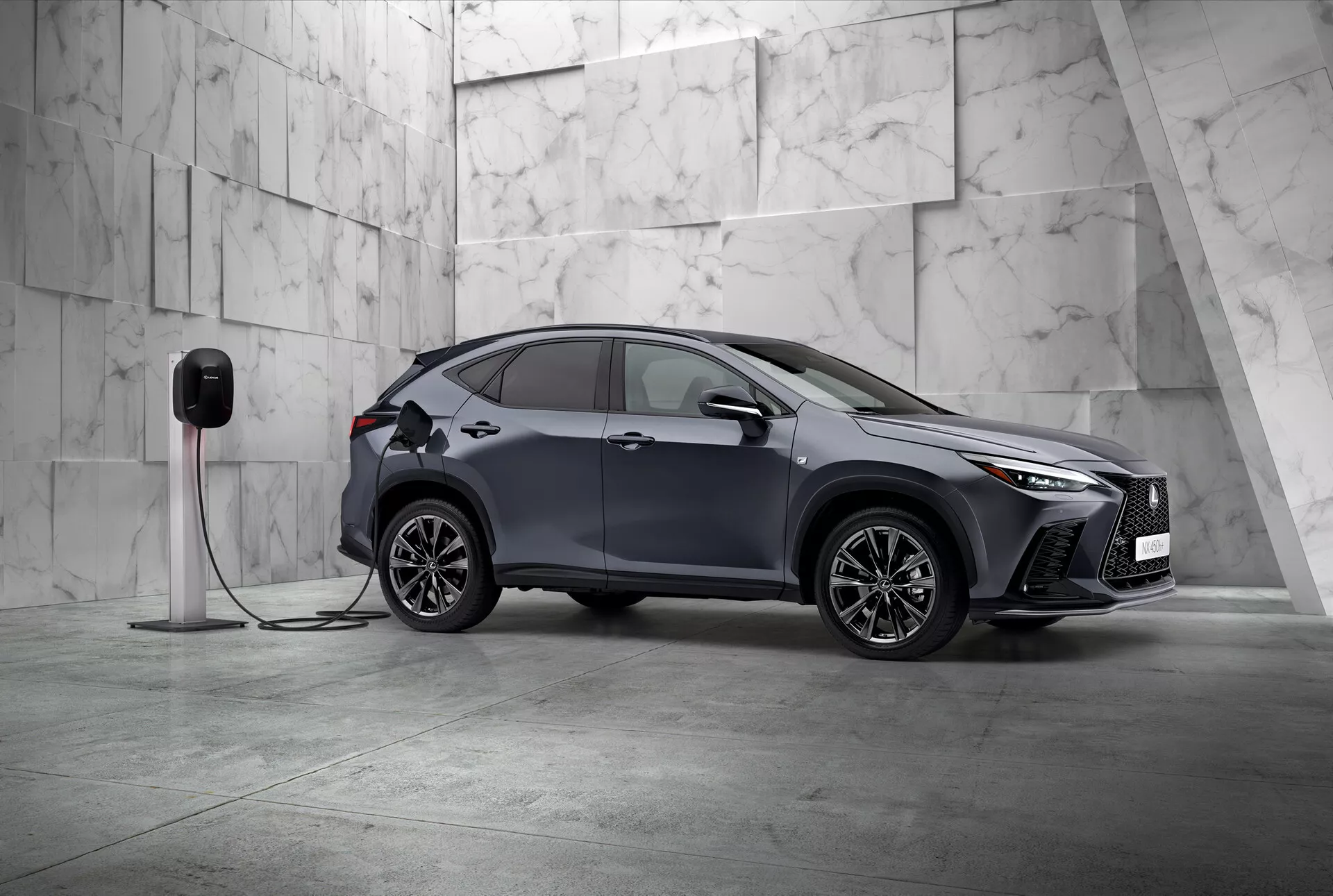Tento obrázek zobrazuje Lexus NX 2021, moderní kompaktní SUV s elegantním a sportovním designem, fotografované z pohledu tři čtvrtiny zepředu. Vidíme levou stranu a částečně i přední část vozidla, umístěného diagonálně v minimalistickém prostředí zdobeném velkými světlými mramorovými dlaždicemi, naznačující prémiovou atmosféru. Lexus NX se může pochlubit výraznými úhlovými LED světly a prominentní přední mřížkou, zdůrazňující jeho sofistikovanost. Viditelné jsou aerodynamicky tvarovaná boční zrcátka, složitá vícelistová slitinová kola a zapuštěné kliky dveří, které zdůrazňují sportovní a pokročilý design. Důležitým detailem je připojení auta k elektrické nabíjecí stanici, což signalizuje, že se jedná o elektrické vozidlo nebo plug-in hybrid. Nabíjecí port na předním blatníku naznačuje jeho elektrické schopnosti. Tento statický snímek se zaměřuje na předvedení designu vozidla a technologických funkcí v klidné, vyvážené kompozici s dostatkem negativního prostoru, což ztělesňuje exkluzivitu a inovaci v automobilovém designu.