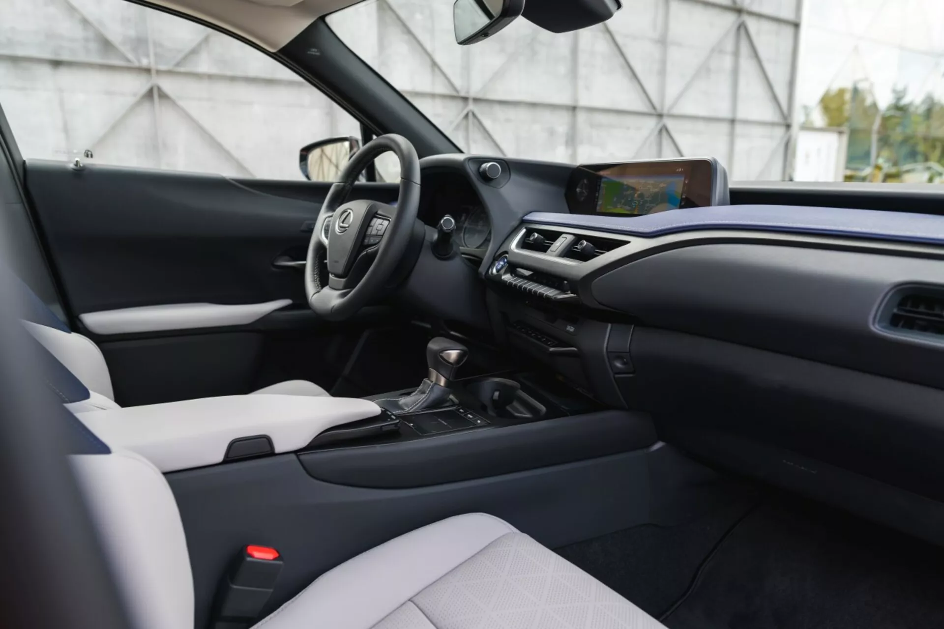 Interiér Lexus UX 2019 z pohledu předního spolujezdce ukazující palubní desku, středovou konzolu, sedadlo řidiče s volantem a digitálními displeji, dotykový informačně-zábavní systém, klimatizaci, volič převodovky, sedadlo spolujezdce čalouněné ve dvoubarevném provedení a moderní dekorační prvky.