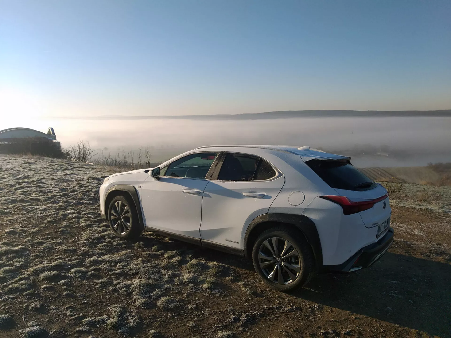 Lexus UX 2019 zaparkovaný venku s zobrazením zadního tříčtvrtečního pohledu zdůrazňujícího jeho aerodynamický design s rychloběžným zavazadlovým prostorem, integrovaným spoilerem, štíhlými horizontálními zadními světly a koly z lehkých slitin s více paprsky.