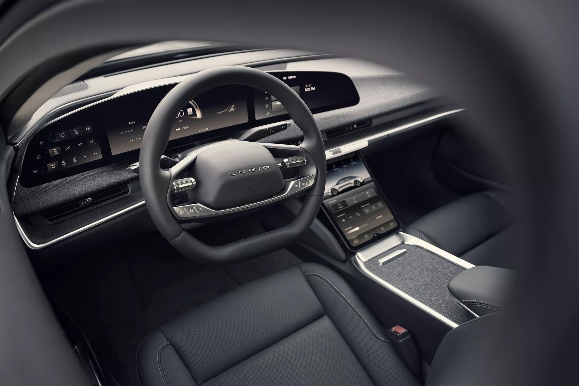 Detailní pohled na kokpit řidiče vozu Lucid Air 2022 z perspektivy zadního sedadla, zobrazující moderní multifunkční volant s ovládacími prvky, široký digitální displej palubní desky, vertikálně orientovanou centrální dotykovou obrazovku s okolními větracími otvory, elegantní středovou konzoli s minimem fyzických tlačítek a luxusní interiér s tmavým barevným schématem obsahujícím vysoce kvalitní materiály.