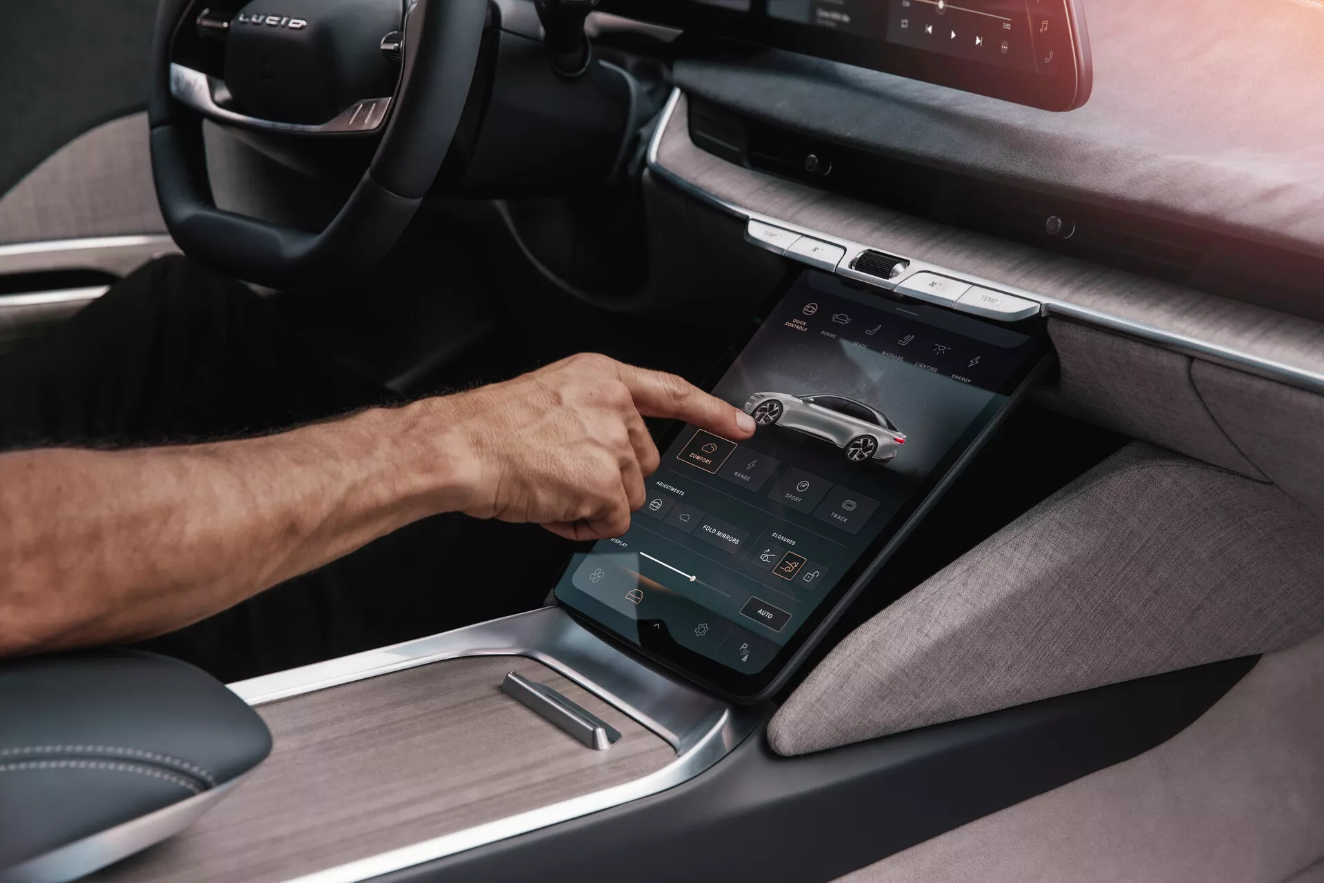 Detailní pohled na středovou konzoli vozu Lucid Air 2022 první generace z šikmého úhlu, zdůrazňující vertikálně orientovaný dotykový displej, na který reaguje pravá ruka osoby, zobrazující pokročilý informační systém s různými funkčními ovladači a kombinací textilního a kovového obložení, naznačující moderní, luxusní interiér zaměřený na technologii.