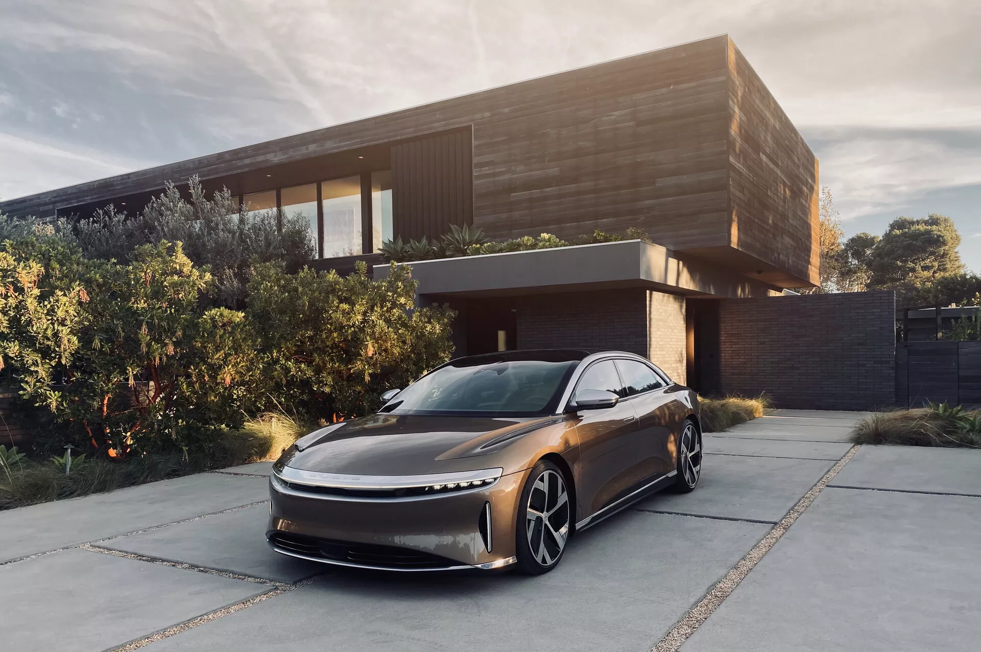 Obrázek sedanu Lucid Air 2022 zaparkovaného před moderním domem. Snímek z nízkého, čelního úhlu tři čtvrtiny, zobrazující jeho elegantní, aerodynamický design s prodlouženou kapotou a subtilní mřížkou. Zvýrazněné jsou tenká LED světla, velká výrazná kola a chybějící boční zrcátka, která naznačují použití kamerového systému zpětného pohledu. Metalická tmavá barva a sofistikované prostředí zvyšují jeho luxusní dojem, což naznačuje, že jde o elektrické vozidlo zaměřené na zámožný trh.