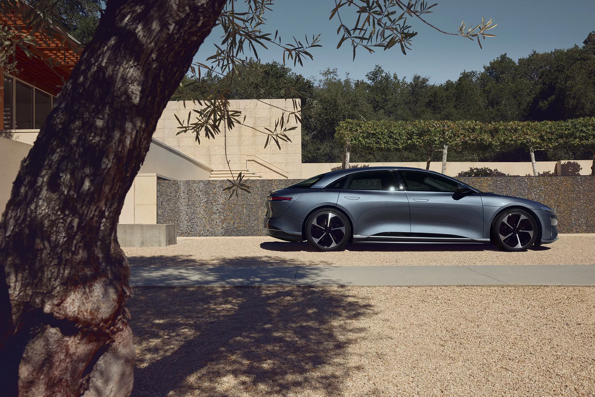 Pohled z profilu na čtyřdveřový sedan Lucid Air 2022 první generace zaparkovaný venku. Viditelnost strany spolujezdce předvádí hladké linie karoserie, design dveří a oken a výrazná hliníková kola s nízkoprofilovými pneumatikami. Částečně zastíněný stromem, s přirozeným světlem zdůrazňujícím kovovou povrchovou úpravu. Moderní budova v pozadí zdůrazňuje elegantní, aerodynamický design vozu s pokročilými funkcemi, jako jsou zapuštěné kliky dveří a aerodynamická zrcátka, která mohou naznačovat integrované směrové světla nebo detekci mrtvého úhlu.