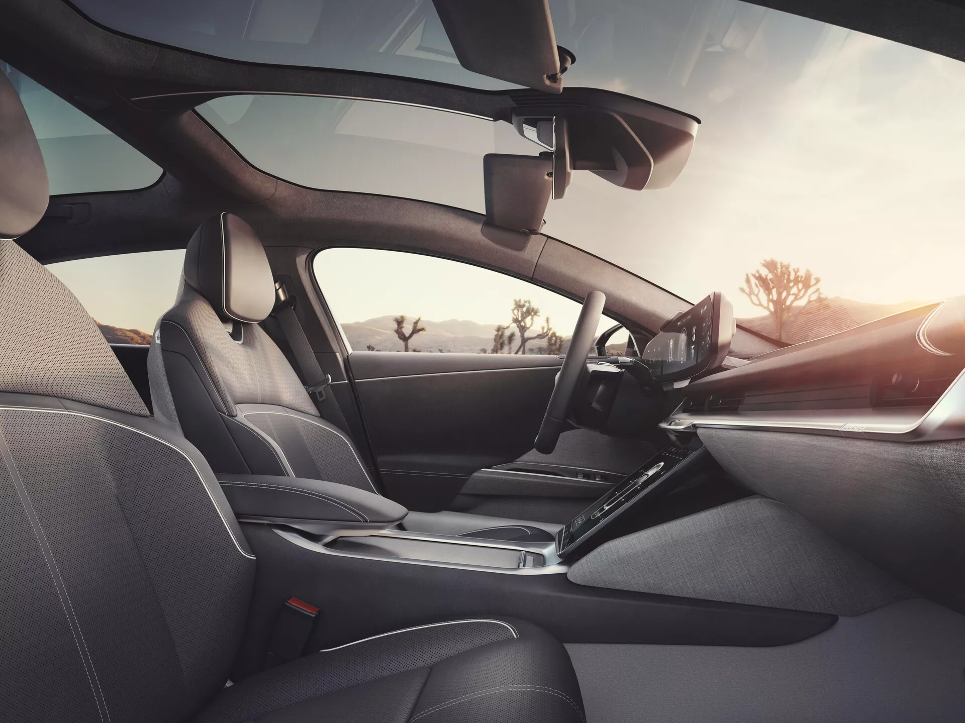 Interiér vozu Lucid Air 2022 (1. generace) z pohledu zadního pasažéra, ukazující sedačky s tmavým koženým čalouněním a detailním prošíváním, elegantní palubní desku s volantem vlevo s integrovanými ovladači, výrazný dotykový informační displej, ergonomickou řadicí páku, dveřní panely s elektronickými ovladači a reproduktory, viditelné zpětné zrcátko a možný asistenční systém řidiče u čelního skla, zdůrazňující luxusní design, pokročilou technologii a pohodlí.