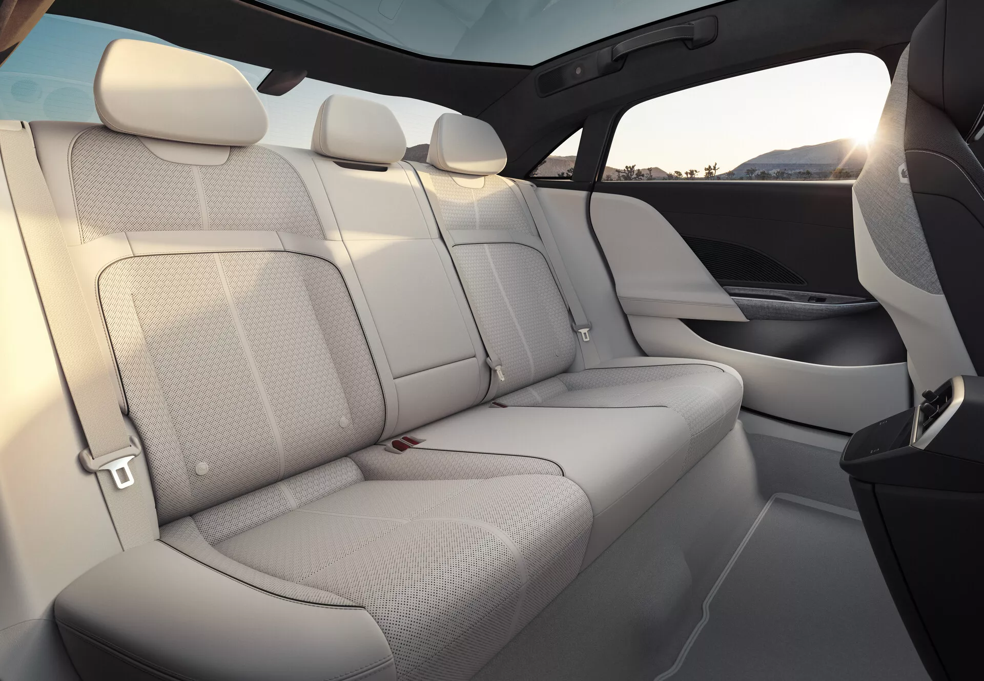 Pohled na interiér vozu Lucid Air 2022, zaměřující se na zadní sedadla pro cestující. Sedadla jsou čalouněna světlou kůží, což interiéru dodává světlý a prostorný pocit. Zadní lavice pojme tři pasažéry s odpovídajícími opěrkami hlavy a viditelnými bezpečnostními pásy. Panely dveří plynule přecházejí s materiálem a designem sedadel, včetně integrovaných mřížek reproduktorů. Vysoká linie oken a rozsáhlá zatmavená panoramatická střecha zvyšují pocit soukromí a ochrany. Interiérový lem ukazuje směs světelné čalounění a tmavších prvků, pravděpodobně z vysoce kvalitních materiálů.