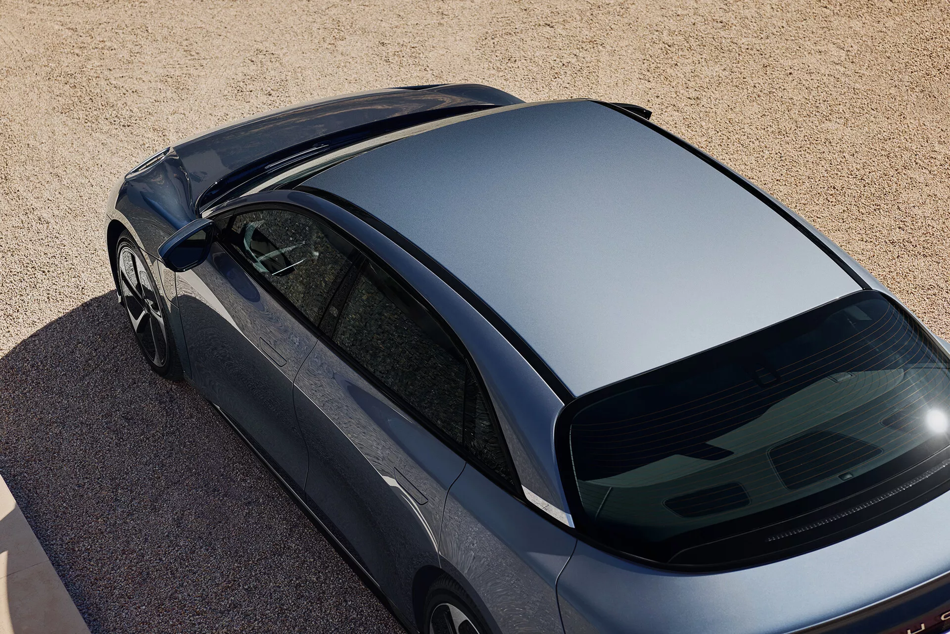 Nadzemní šikmý pohled na tmavě kovový Lucid Air 2022 1. generace, zaparkovaný na povrchu ze štěrku nebo hrubého písku. Snímek zachycuje pohled shora se zaměřením na aerodynamickou střechu, prudce sklopené zadní okno a část kufru. Vlastnosti zahrnují bezrámová okna, aerodynamická boční zrcátka a absenci střešních doplňků a zadního stěrače, což naznačuje aerodynamický design a moderní styl.