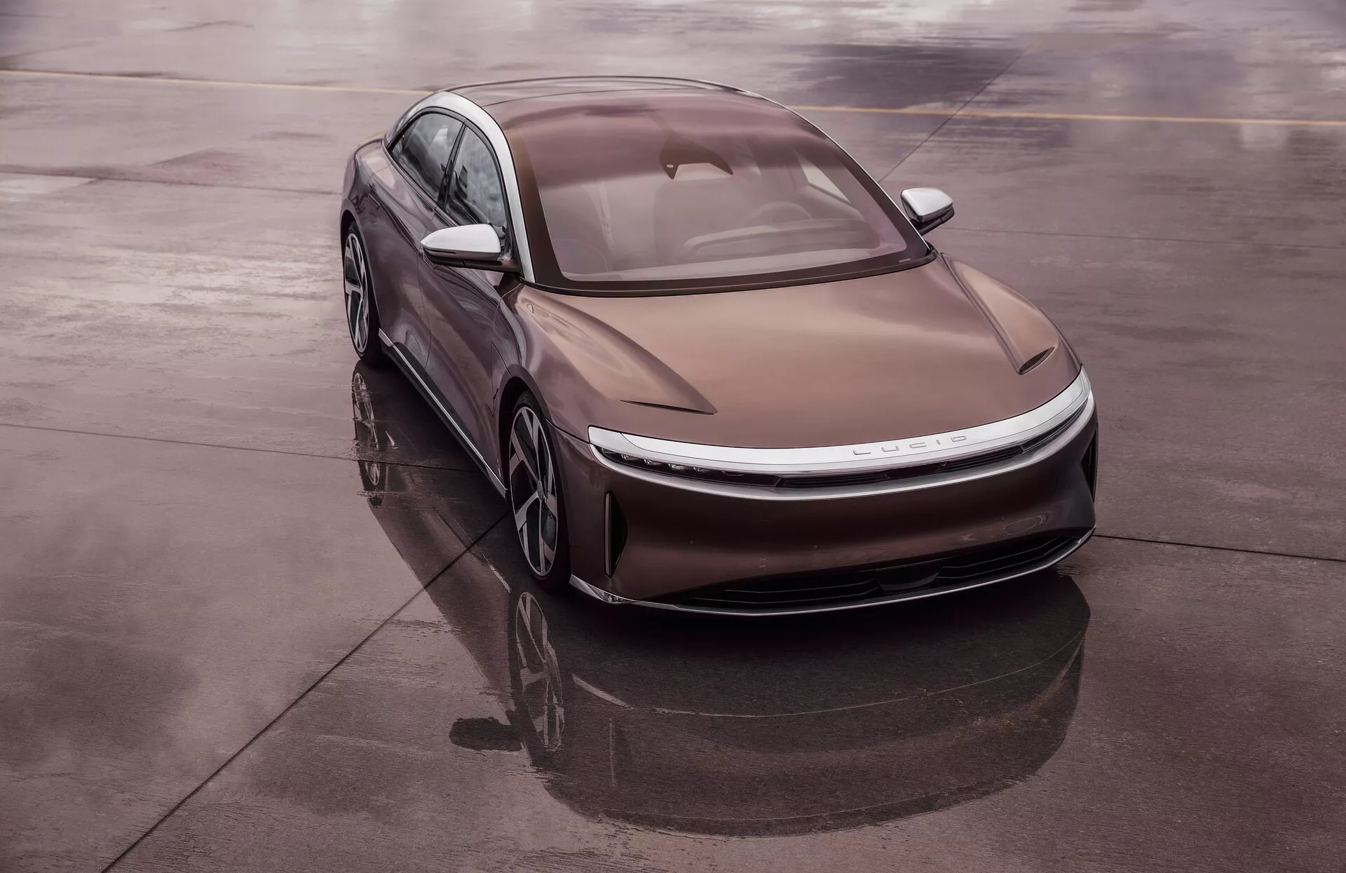 Vysokoúhlý snímek moderního sedanu Lucid Air 2022, 1. generace, diagonálně umístěného na reflexním povrchu, jako je mokrý asfalt, poskytuje komplexní pohled na přední levý čtvrtletní vůz. Elegantní, aerodynamický design obsahuje hladké křivky, tenké LED světlomety a tvarovanou kapotu s výraznými liniemi. Střecha připomínající fastback a propláchnuté sklo zlepšují aerodynamickou účinnost. Elektrický charakter vozu naznačuje absence tradiční mřížky, integrovaného sacího systému a lehkých aerodynamických kol.