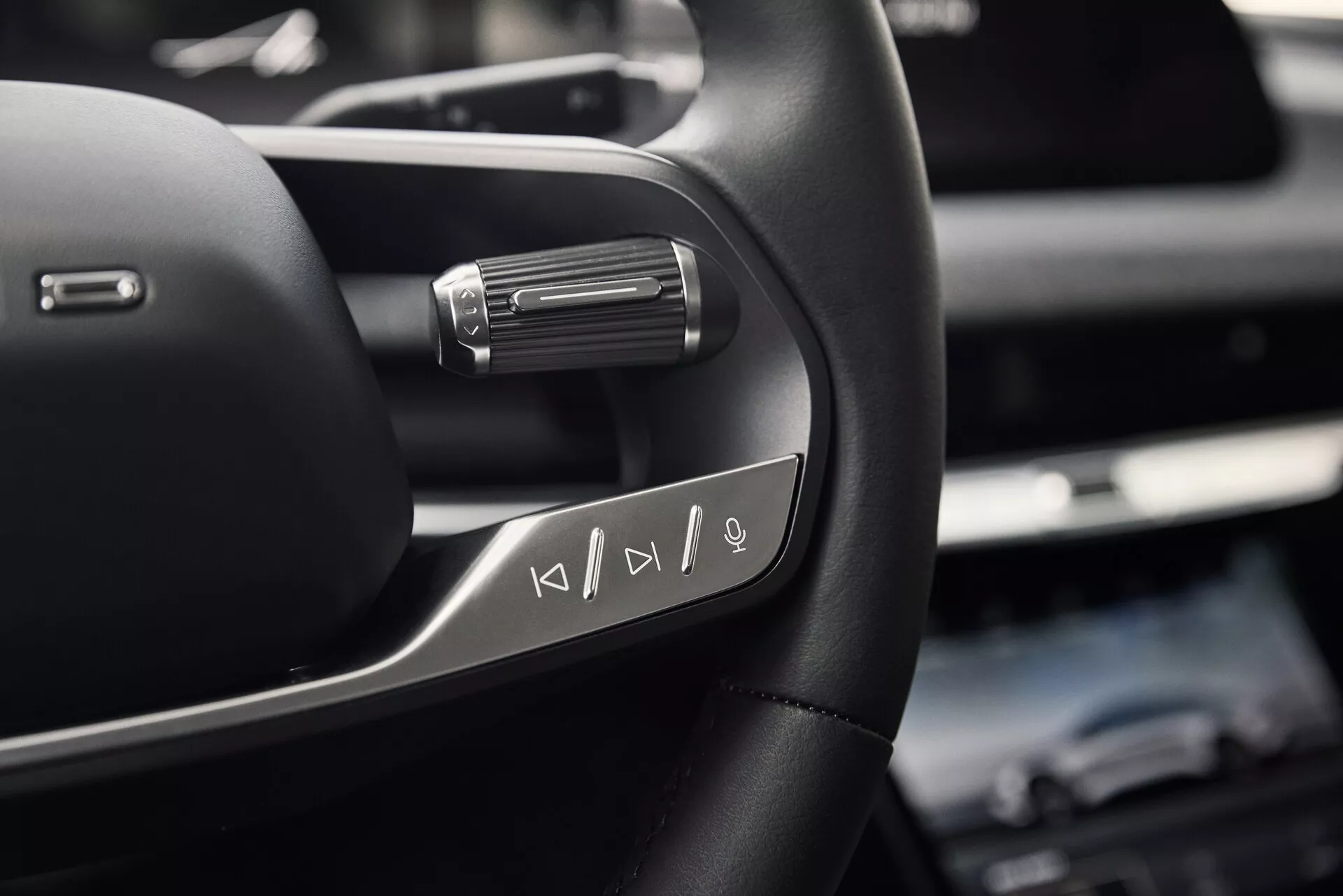 Detailní pohled na levou stranu volantu Lucid Air 2022 s multifunkčními tlačítky pro ovládání médií. Fotografie pořízená zespodu volantu na úrovni hrudi sedícího řidiče zdůrazňuje vysoce kvalitní kožený materiál s kovovými akcenty a pádlem za volantem, což naznačuje pokročilé jízdní funkce.