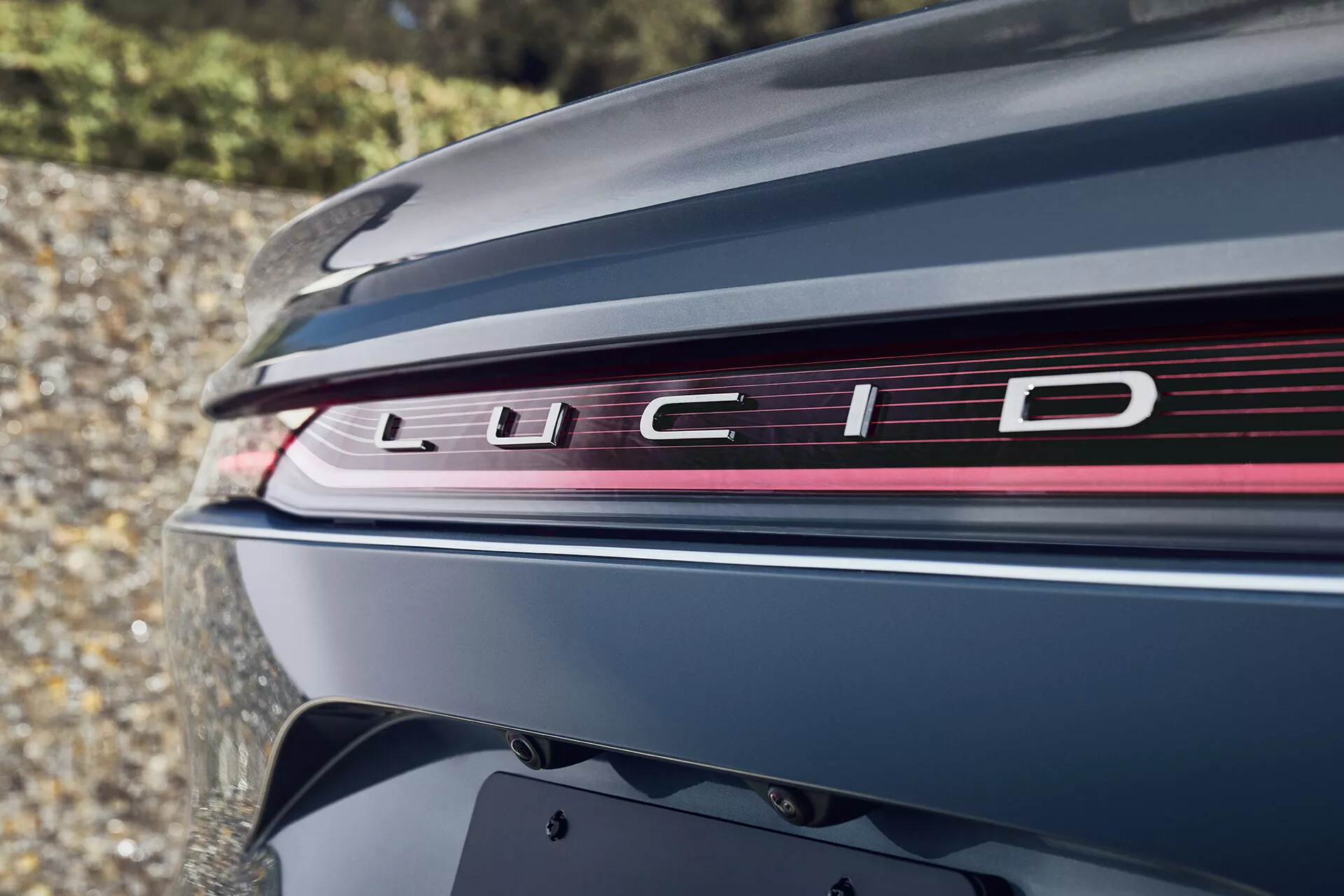 Detailní záběr zadní části vozu Lucid Air 2022 (1. generace) v hluboké metalické barvě. Obrázek zvýrazňuje víko kufru s nápisem 'LUCID' a elegantní světelnou lištu přes celou šířku, integrující brzdová světla a směrovky. Mírně nízký úhel zdůrazňuje aerodynamický design vozu, vysoce kvalitní lak a nejmodernější automobilovou technologii, která je synonymem pro luxusní elektromobily.