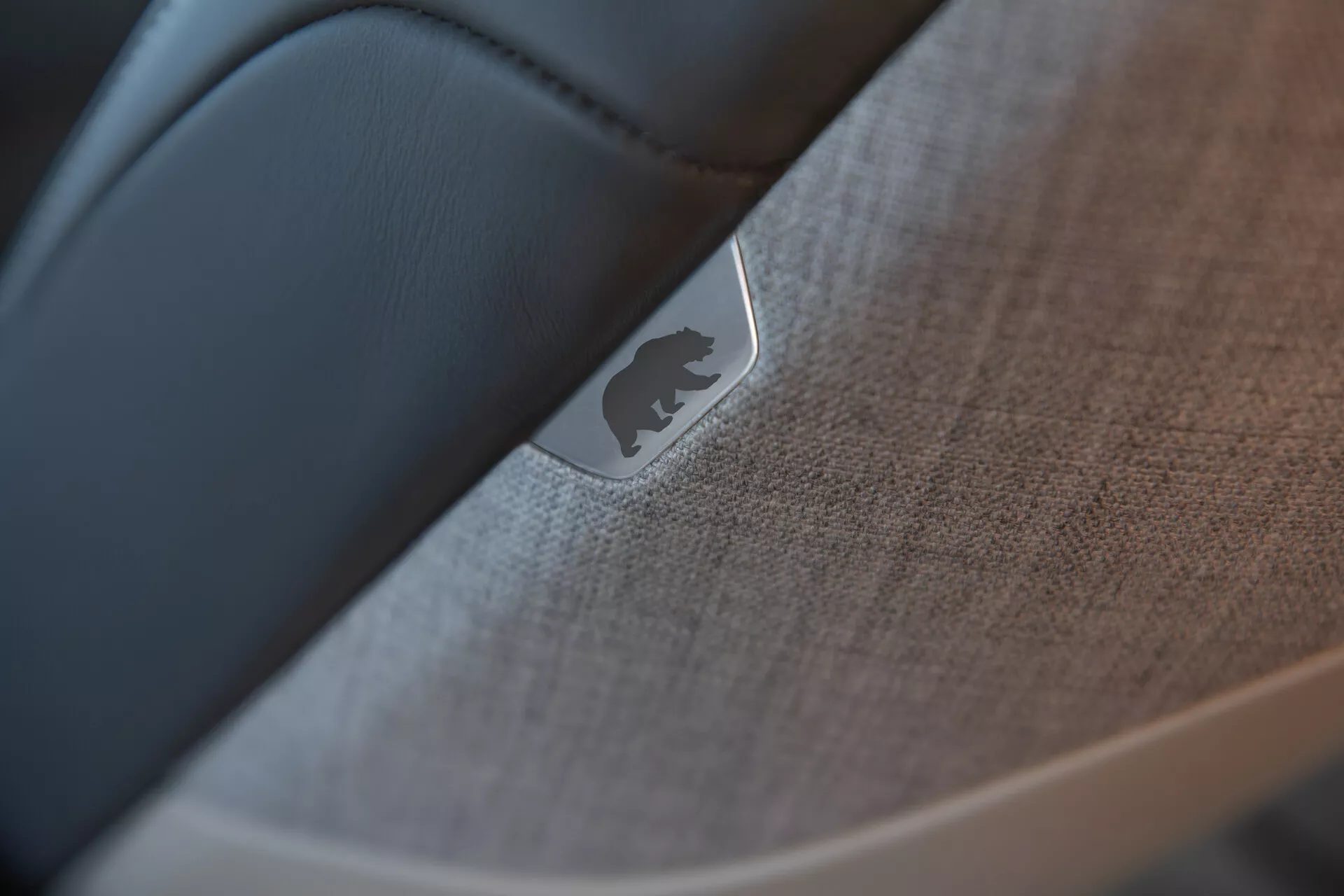 Detailní záběr, selektivní zaostření na interiér vozu Lucid Air 2022 (1. generace) zdůrazňující detail dveří nebo palubní desky. Reflektivní kovový emblém nebo odznak je prominentně umístěn na texturované látce nebo měkkém plastu s detaily šití. Hladký, tmavší materiál, pravděpodobně černá kůže, kontrastuje s texturovanými prvky, ukazující řemeslné zpracování a vysokou kvalitu interiéru vozu.