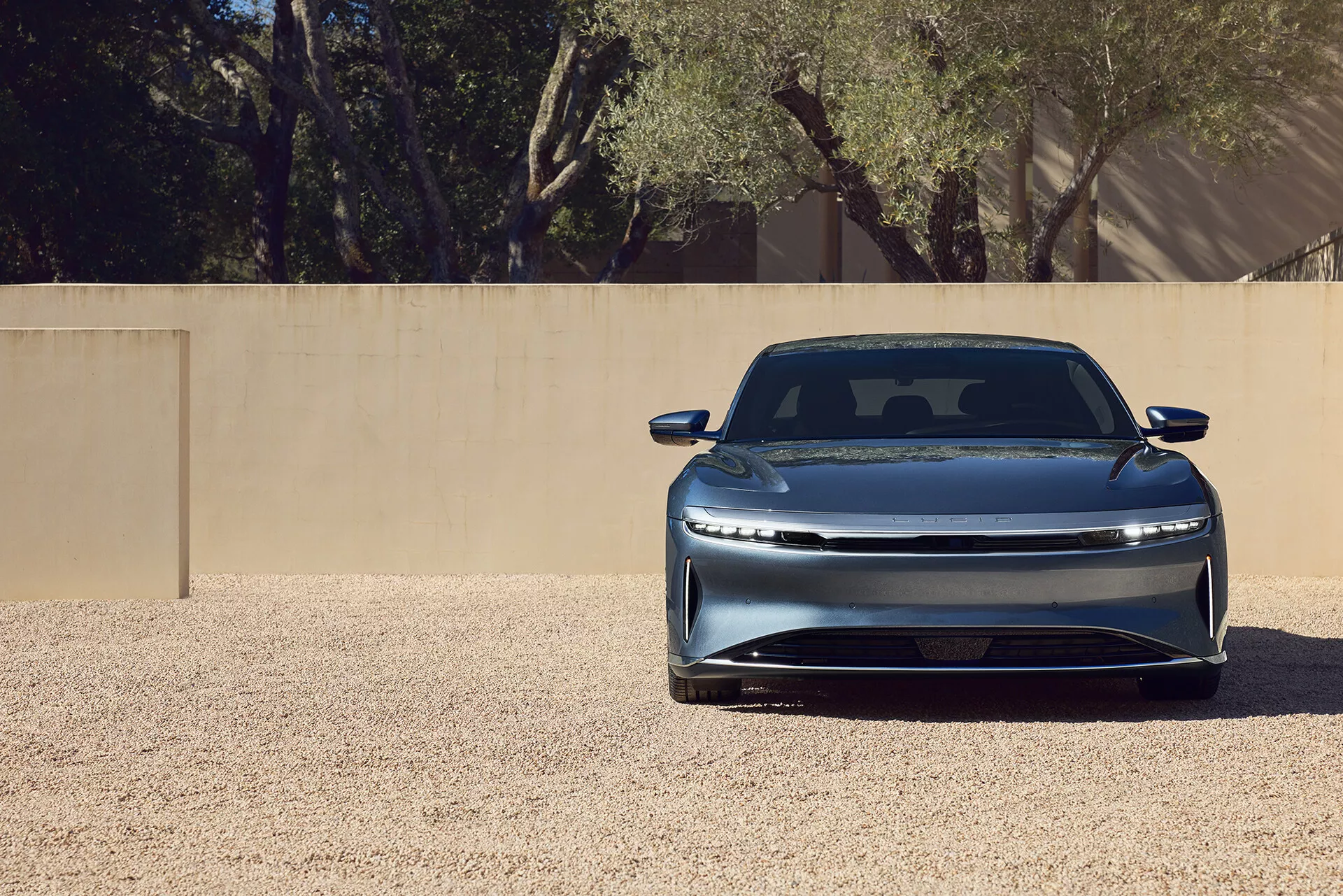 Detailní pohled zepředu na elektrické vozidlo Lucid Air 2022 první generace, zdůrazňující jeho elegantní uzavřenou masku, horizontálně orientovaná LED světla, integrovaný přední nárazník, tvarovanou kapotu s obrysovou linií a metalický lak. Vozidlo je centrálně umístěno na štěrkovém povrchu pod jasným osvětlením, což naznačuje pokročilou aerodynamiku a moderní luxusní design.