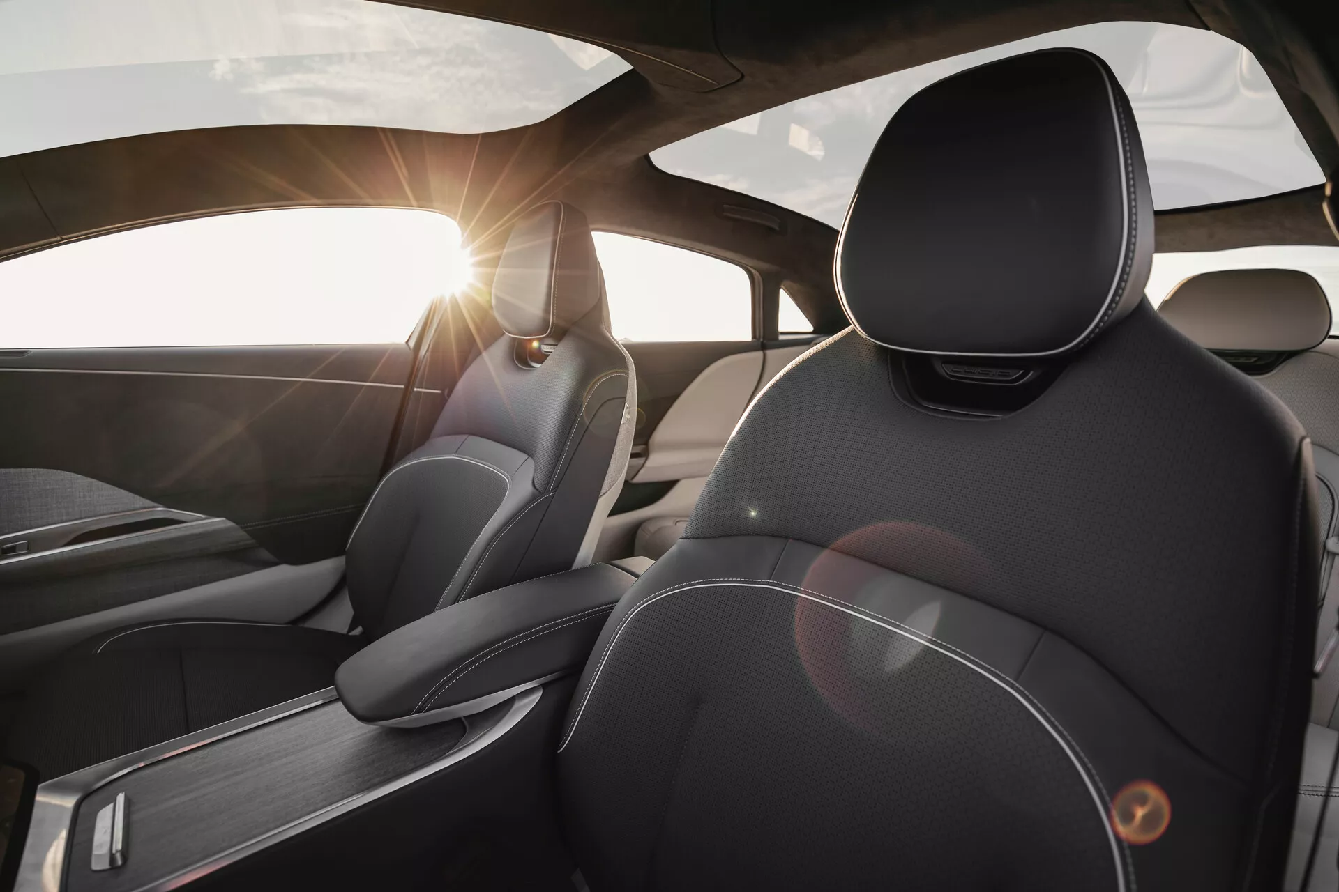 Pohled na interiér modelu Lucid Air 2022 z pohledu zadního sedadla, zaměřený na řidičovo sedadlo. Zdůrazňuje prémiové materiály a design, včetně detailního prošívání na koženém čalounění, ergonomicky navržených opěrek hlavy, kartáčovaných kovových akcentů a světelného efektu od slunce prosvítajícího čelním sklem.