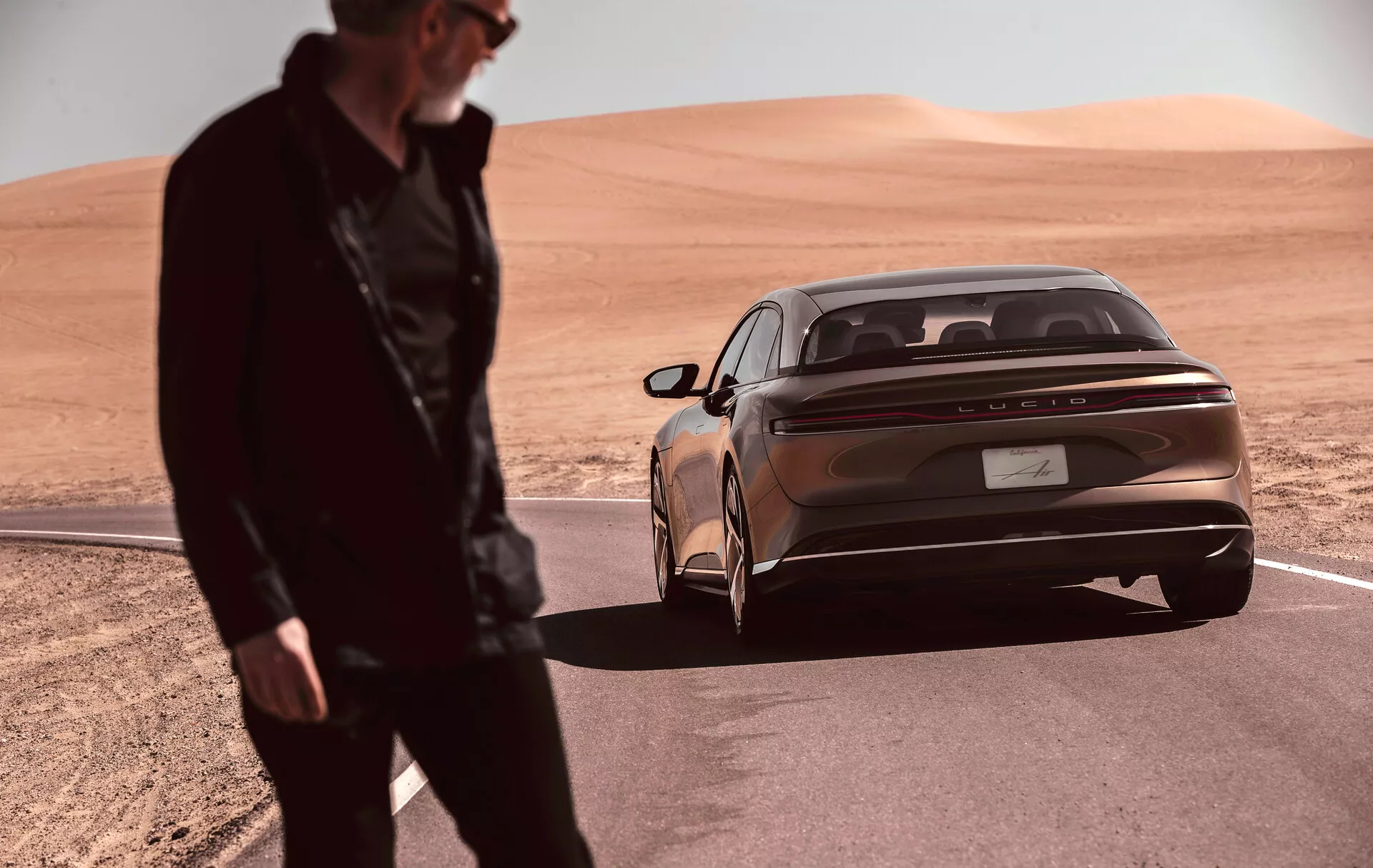 Elegantní a moderní sedan Lucid Air 2022 (1. generace) zaparkovaný v pouštní krajině, zachycený z nízkého úhlu s důrazem na zadní tři čtvrtiny vozu. Kovová bronzová barva karoserie a výrazná horizontální zadní světla podtrhují současný design vozu. Pod světly prochází reflexní chromovaný pás. Na kufru je centrálně umístěna značka výrobce. Integrovaný zadní nárazník je vybaven parkovacími senzory, což přidává na luxusním vzhledu. V popředí stojí rozmazaná postava, naznačující pohyb nebo aktivitu v blízkosti vozidla.