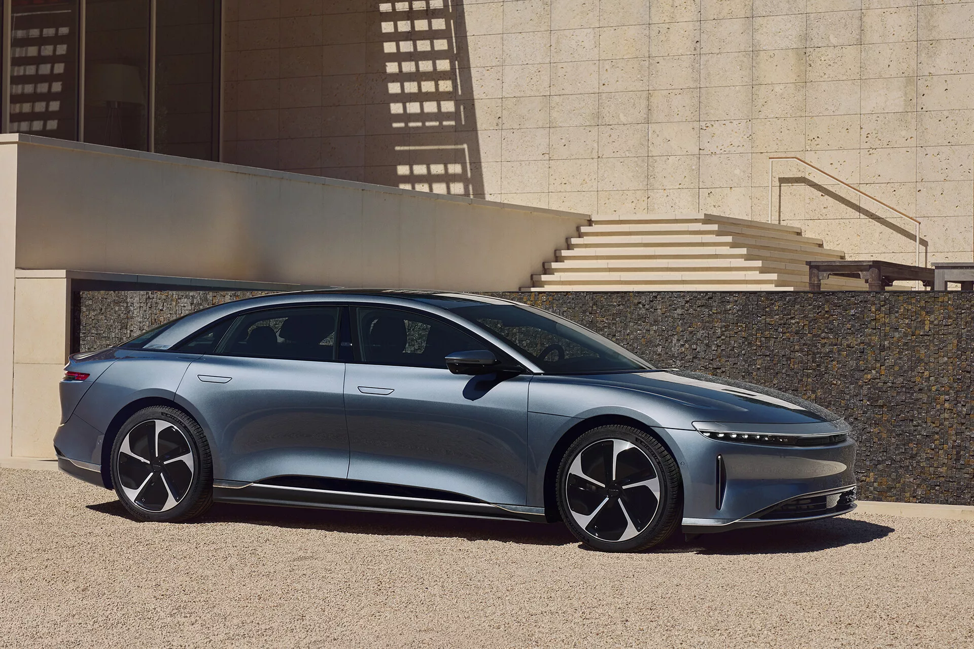Boční profil vozu Lucid Air 2022, moderního elegantního sedanu, zachycený venku v městském prostředí s architektonickými prvky vrhajícími stíny. Levá strana vozu ukazuje jeho elegantní, aerodynamický design s nízkou střechou, zapuštěnými klikami dveří, velkými vícepaprskovými koly a úzkými bočními zrcátky. Obrázek zvýrazňuje délku vozu od předního po zadní nárazník, raked-forward A-sloupek pro vzhled prostorného interiéru a dynamickou charakterovou linií na karoserii.