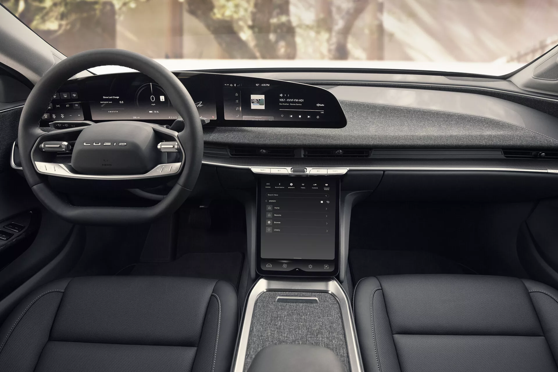 Pohled ze zadního středového sedadla interiéru Lucid Air 2022, který ukazuje moderní design s koženým multifunkčním volantem, širokým digitálním displejem na palubní desce, vertikálně orientovanou dotykovou obrazovkou na středové konzoli, pákami pro manuální výběr převodů, elegantními koženými a textilními předními sedadly, středovým tunelem s řadicí pákou a držáky na nápoje a dveřními panely s úložnými kapsami a mřížkami reproduktorů.
