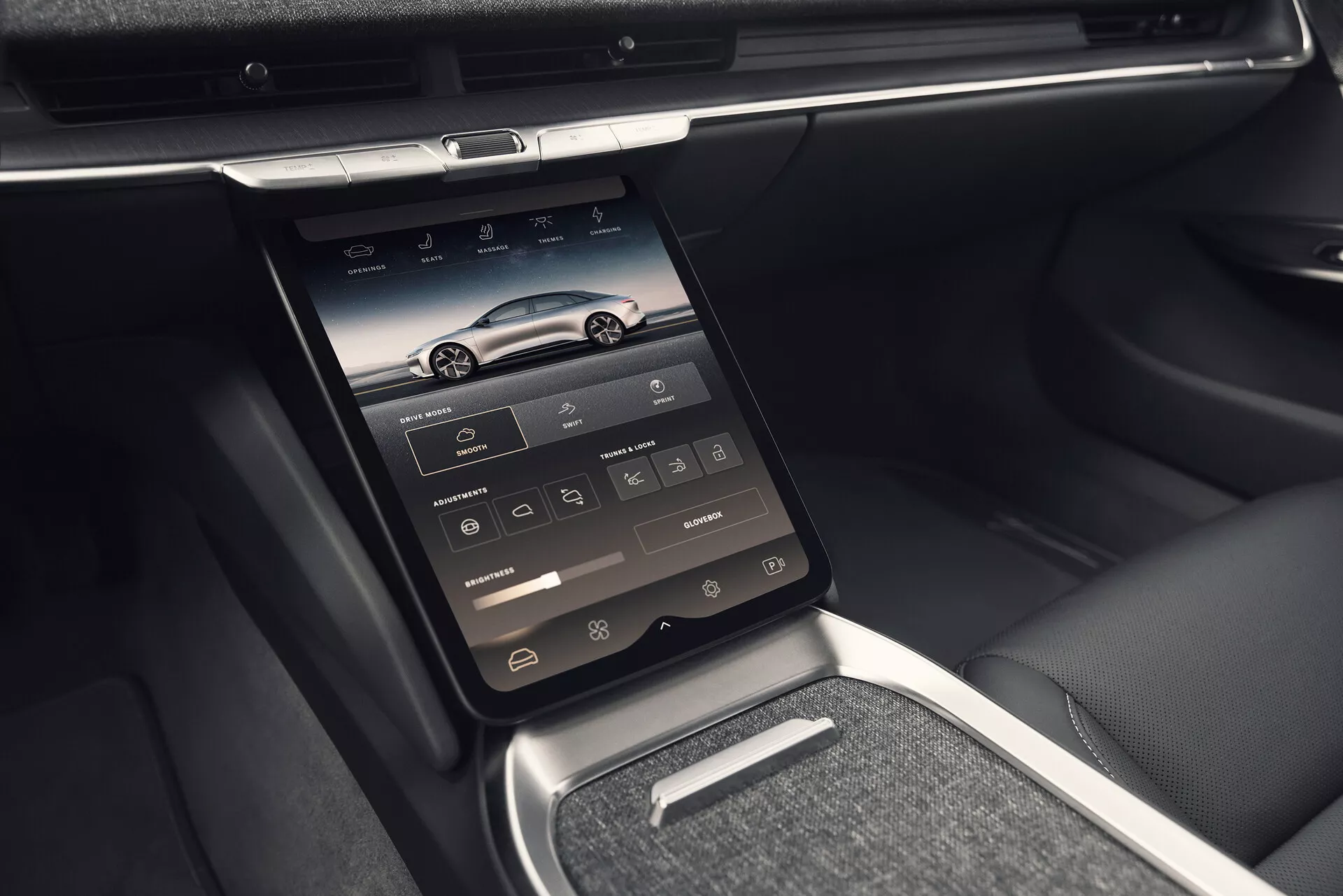 Pohled do interiéru Lucid Air 2022 (1. generace) představujícího středovou konzoli s vertikálně umístěným dotykovým displejem, na kterém jsou ikony navigace, telefonu, médií, klimatizace a nastavení vozidla. Fotografie pořízena z mírně levého úhlu, zvýrazňující pokročilé technologické funkce s ventilačními otvory nad obrazovkou, lesklými chromovanými nebo kovovými ozdobnými prvky, elegantní přístrojovou deskou a směsí kůže a kovových povrchů. Obrázek také ukazuje malou polici pod dotykovou obrazovkou, kovový volič převodovky a kůží potažené opěrky a boky sedadel, zdůrazňující luxusní a pohodlný interiér.