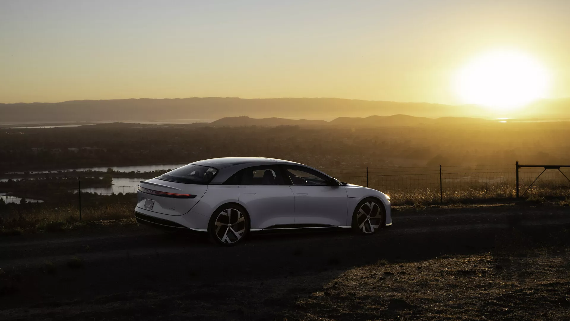 Lucid Air 2022 (1. generace) fotografovaný z úhlu zezadu ze strany za svítání nebo soumraku s teplým sluncem na pozadí, umístěný na levé straně záběru. Elegantní, moderní sedan se čtyřmi dveřmi má hladké linie, velká kola s více paprsky a zatahovací dveřní kliky. Krajina zahrnuje vodní plochy a vysokou vyhlídku. Zadní světla jsou úzká a horizontálně protažená, sluneční světlo vrhá na scénu teplou záři a efekt lens flare.
