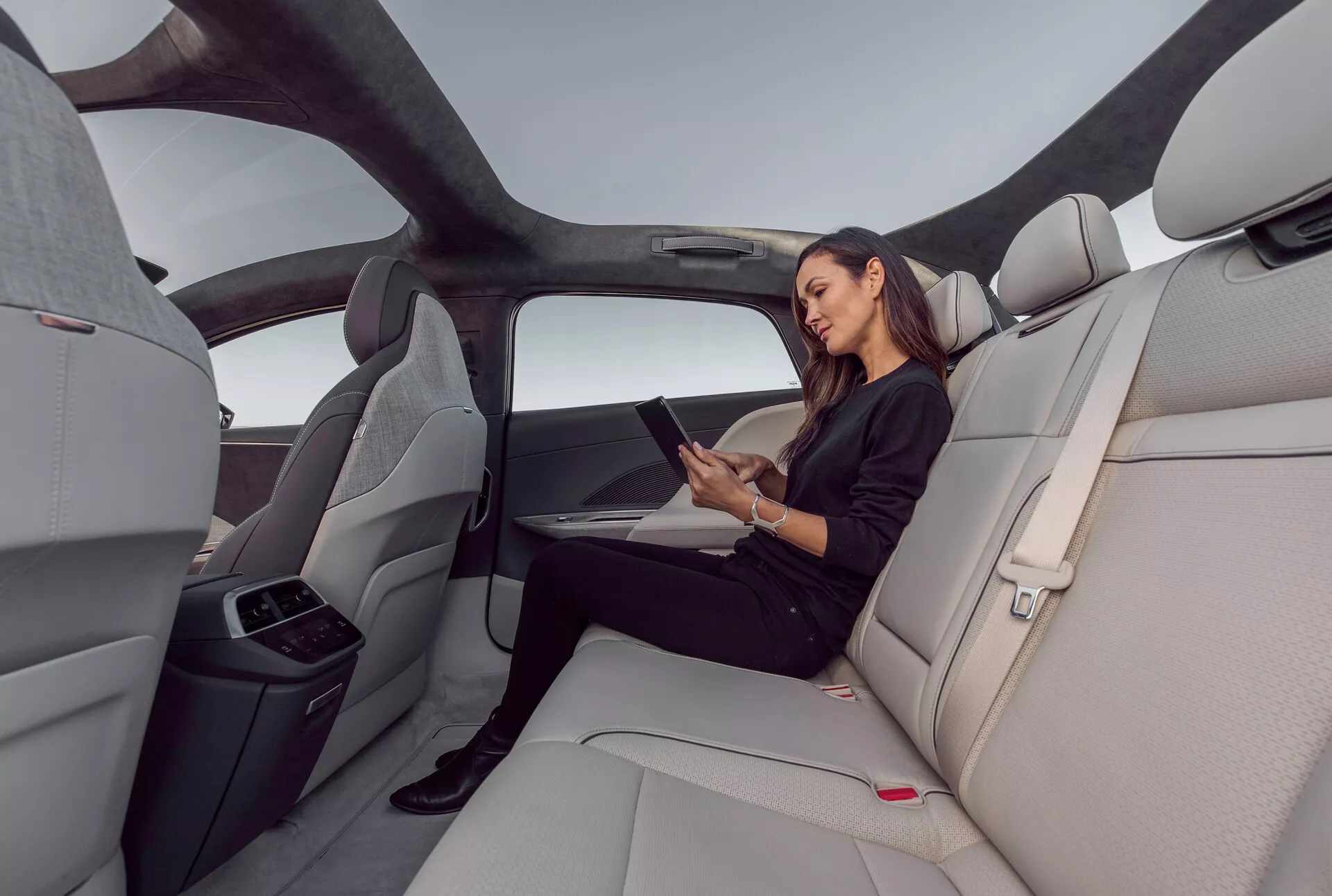 Pohled na interiér zadní části vozu Lucid Air 2022, zdůrazňující prostornou, luxusní kabinu s světle šedými, tvarovanými koženými sedadly. Obrázek zachycuje širokoúhlý záběr z pravé strany, představující panoramatickou střechu, která se táhne přes zadní prostor, ergonomicky navržená sedadla a sklopenou střední opěrku rukou s držáky na nápoje. Žena sedí pohodlně a používá tablet, což naznačuje možnost připojení k informačním technologiím. Elegantní, minimalistický interiér s červenými tlačítky na bezpečnostních pásech a vodorovnými liniemi na palubní desce a dveřních panelech, které zvyšují pocit šířky a otevřenosti.