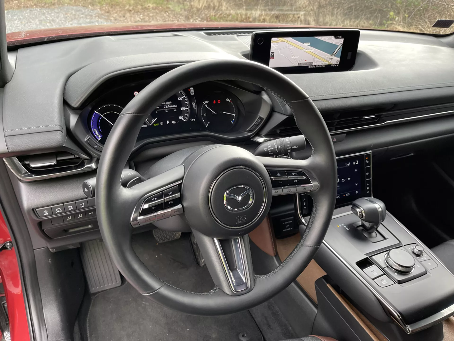 Interiér Mazda MX-30 2020 z pohledu řidiče se zaměřením na detaily jako je volant obalený kůží s třemi větvemi s ovládacími tlačítky a pádly pro řazení, digitální panel přístrojů na palubní desce s modrými akcenty, dotykový informačně-zábavní systém s navigací na centrální konzole, sportovně vyhlížející páka řazení, otočný volič režimu jízdy, další ovládací knoflík informačně-zábavního systému, luxusní kombinace materiálů s černým a červeným lemem, držák na nápoje a ovládací panely na dveřích pro boční zrcátka a okna, které ukazují moderní technologii a ergonomický design.