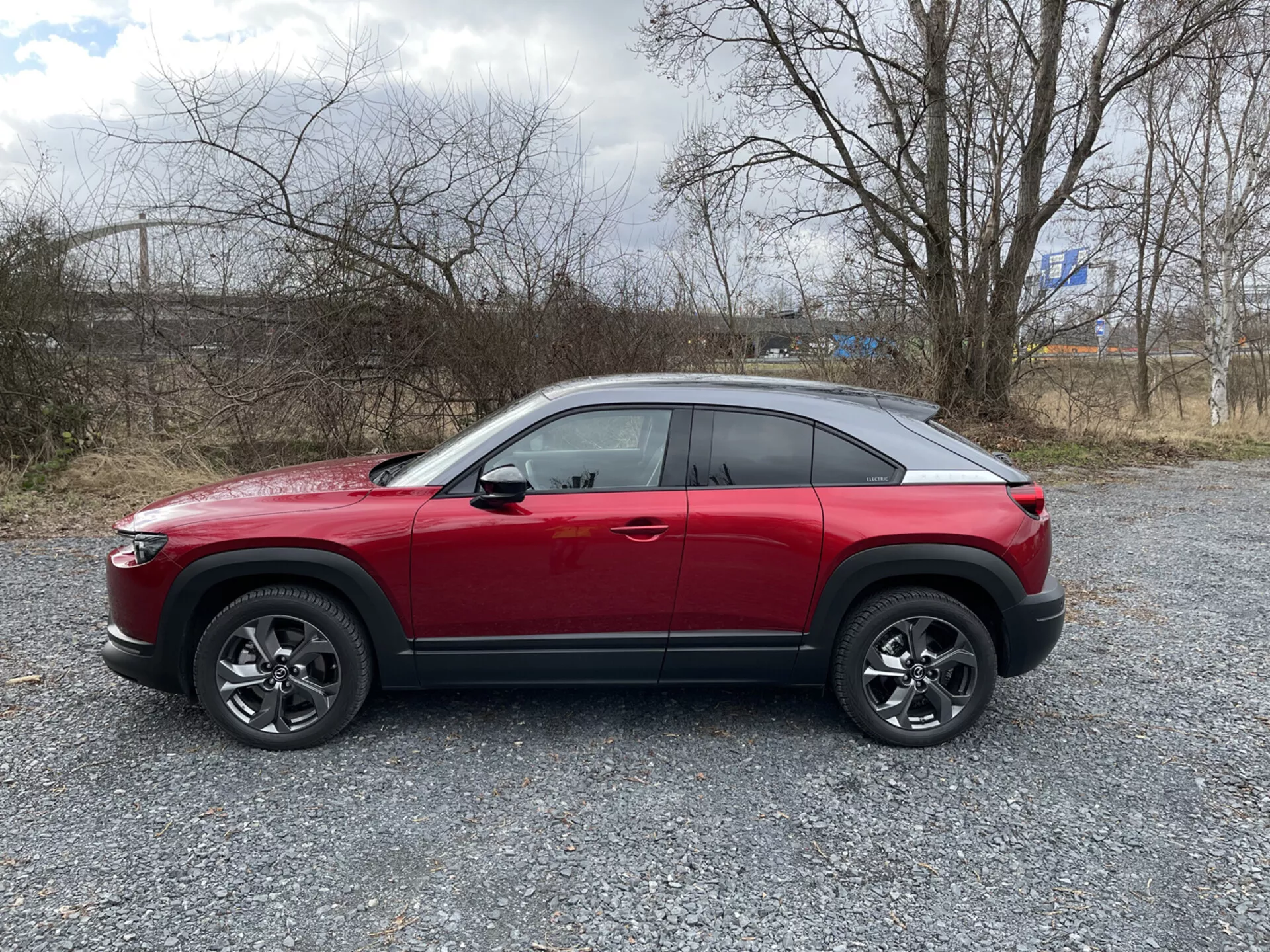 Boční profil červeného crossover SUV Mazda MX-30 z roku 2020 na štěrku s řídkou vegetací a zamračeným nebem, předvádí vysokou linii pásu, kupé-podobnou svažující se střechu, dynamickou charakterovou linii, vícepaprsková slitinová kola s černým obložením a možné pokročilé technologické funkce jako ADAS, LED osvětlení, bezklíčový přístup, TPMS a informačně-zábavní systém.