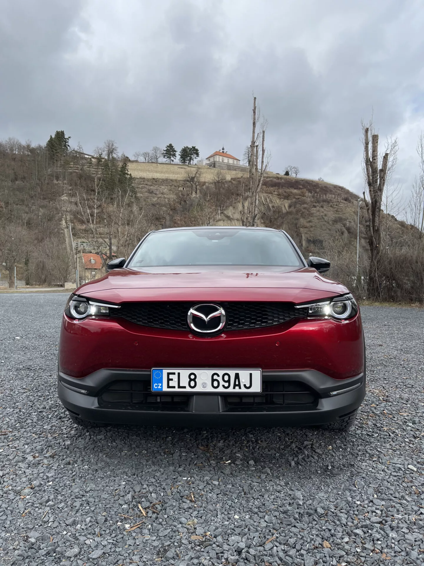 Přední pohled na červené Mazda MX-30 z roku 2020, zdůrazňující mřížku, nárazník a LED světlomety vozu na štěrkovém povrchu proti pozadí zamračené oblohy s zelení a budovou