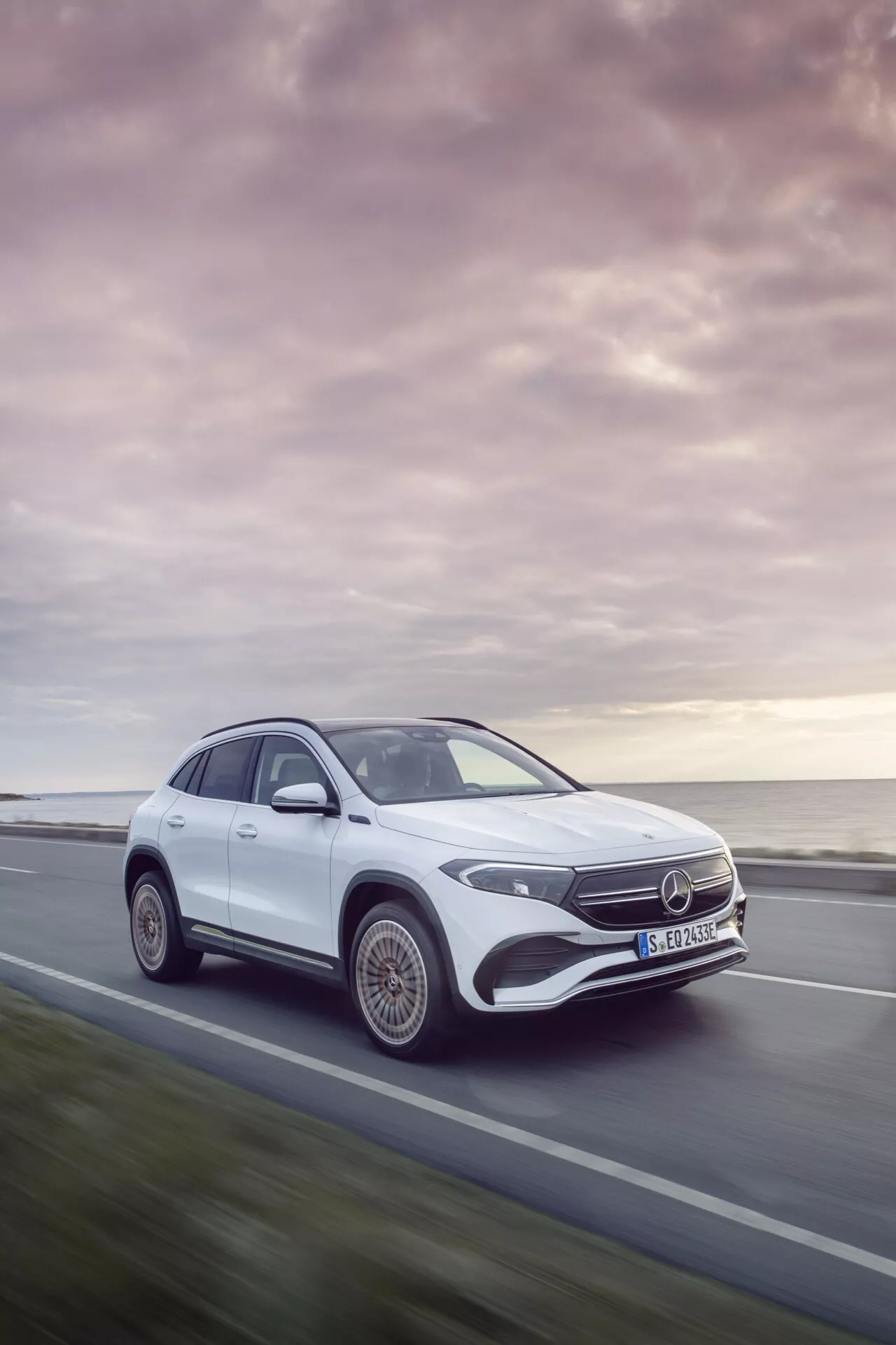 Mercedes-Benz EQA 2021 v pohybu, zobrazený z dynamického tříčtvrtečního předního úhlu představujícího design vozidla, LED světlomety, stylová slitinová kola a aerodynamický siluetu na pozadí klidného západu slunce
