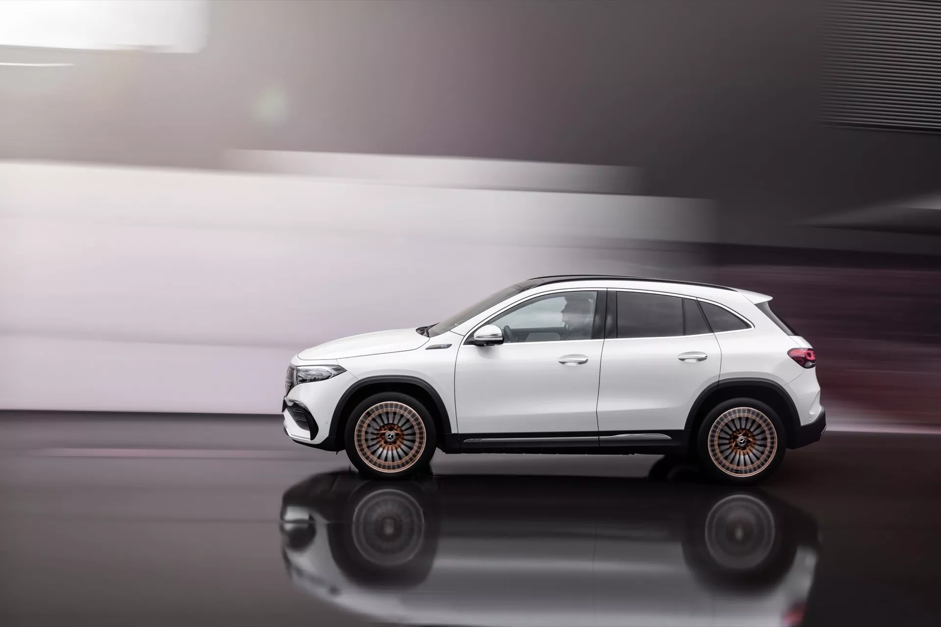 Dinamický profil z boku Mercedes-Benz EQA 2021 v pohybu na leštěné podlaze s vyzdvihnutím kol s vícepaprskovým designem, složených zrcátek, střešních ližin a tvarovaných linií karoserie s okny s ochranným tónováním, předvádějící terénní postoj a potenciální technologické funkce jako automatické sklápění zrcátek a satelitní anténu.