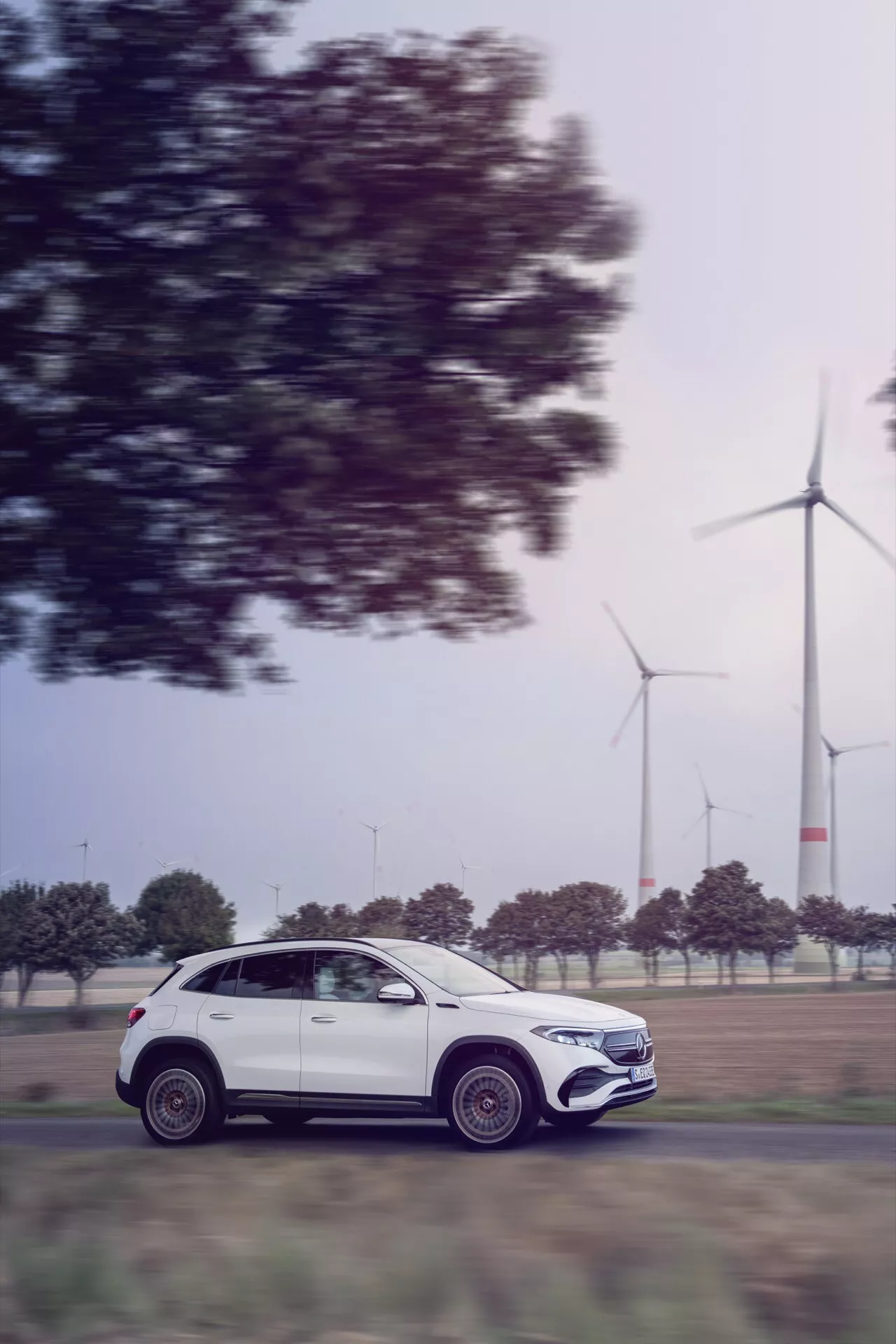 Mercedes-Benz EQA 2021 v pohybu, boční profil zobrazující luxusní design a aerodynamiku, s větrnými turbínami na pozadí naznačujícími ekologickou technologii