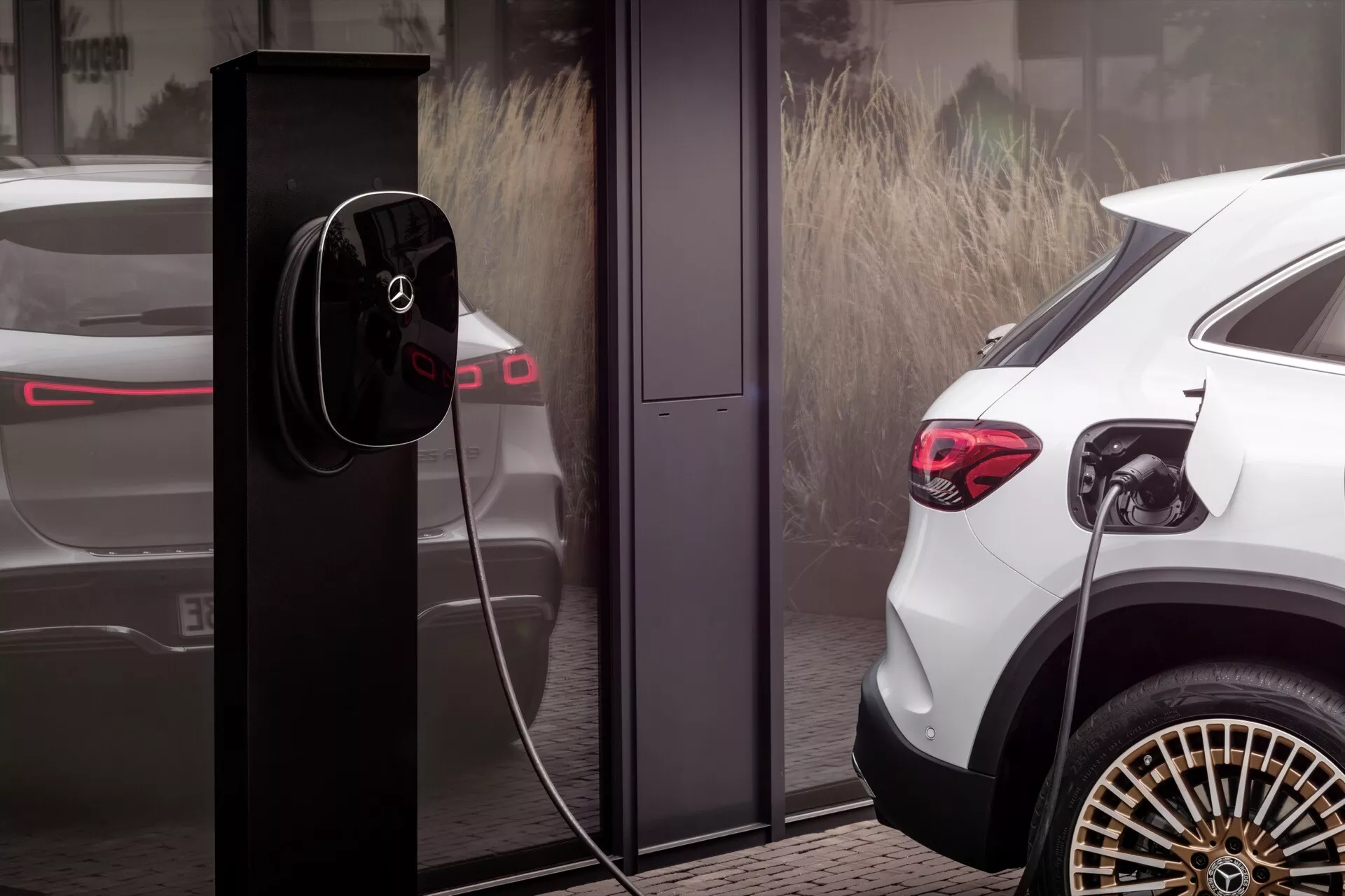 Bílé SUV Mercedes-Benz EQA z roku 2021 zobrazené z úhlu zadního tříčtvrtečního pohledu, které zdůrazňuje jeho vysokou světlou výšku, výrazná LED zadní světla, stylová kola z lehkých slitin s vícepaprskovým designem a připojené k moderní stanici pro nabíjení elektromobilů