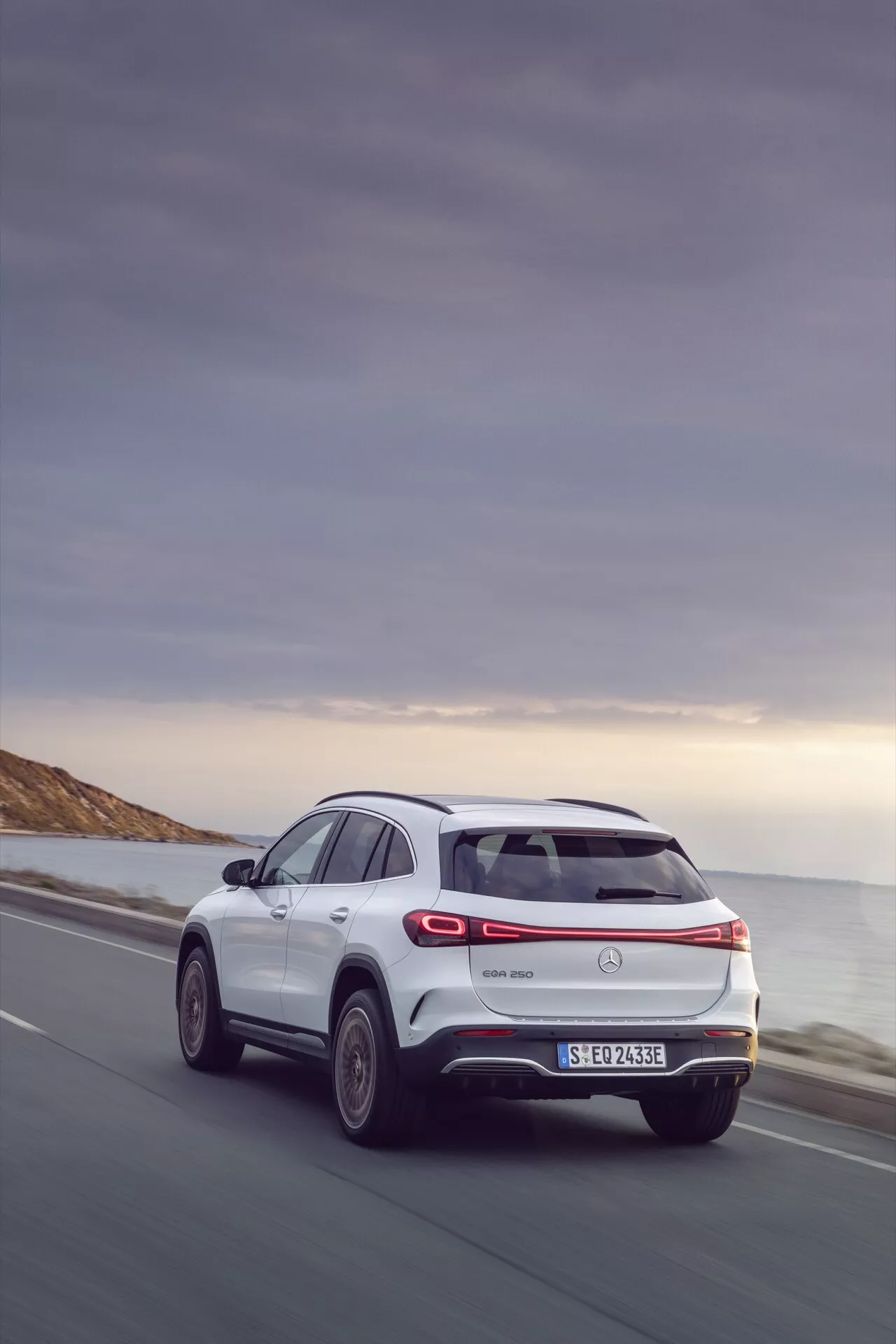 Zadní tříčtvrteční pohled na Mercedes-Benz EQA 243 z roku 2021 na pobřežní silnici, zdůrazňující boční profil, zadní design s LED zadními světly, logo výrobce, zadní stěrač, nárazník s integrovanými odrazkami a parkovacími senzory, dekorativní výfuk a indikace elektrického modelu, velká kola s designem z lehkých slitin s více paprsky, naznačující SUV s potenciální pokročilou aerodynamikou a schopnostmi pohonu všech kol pod večerní oblohou