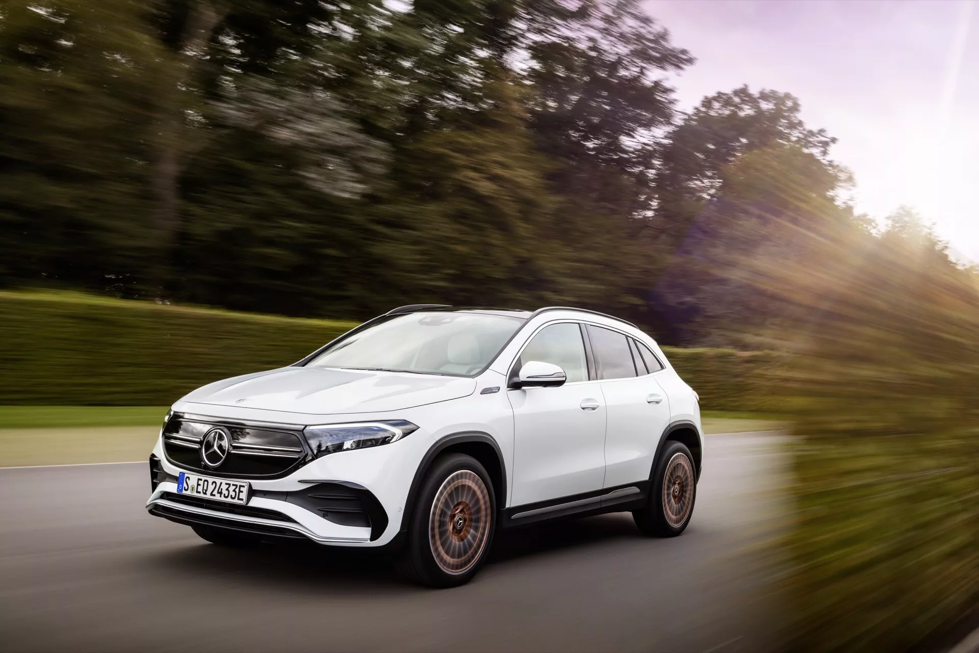 Bílý SUV Mercedes-Benz EQA 2021 v pohybu, pohled z předního tříčtvrtečního úhlu zdůrazňující dynamický postoj, aerodynamický design, LED světlomety a charakteristická slitinová kola na malebné silnici