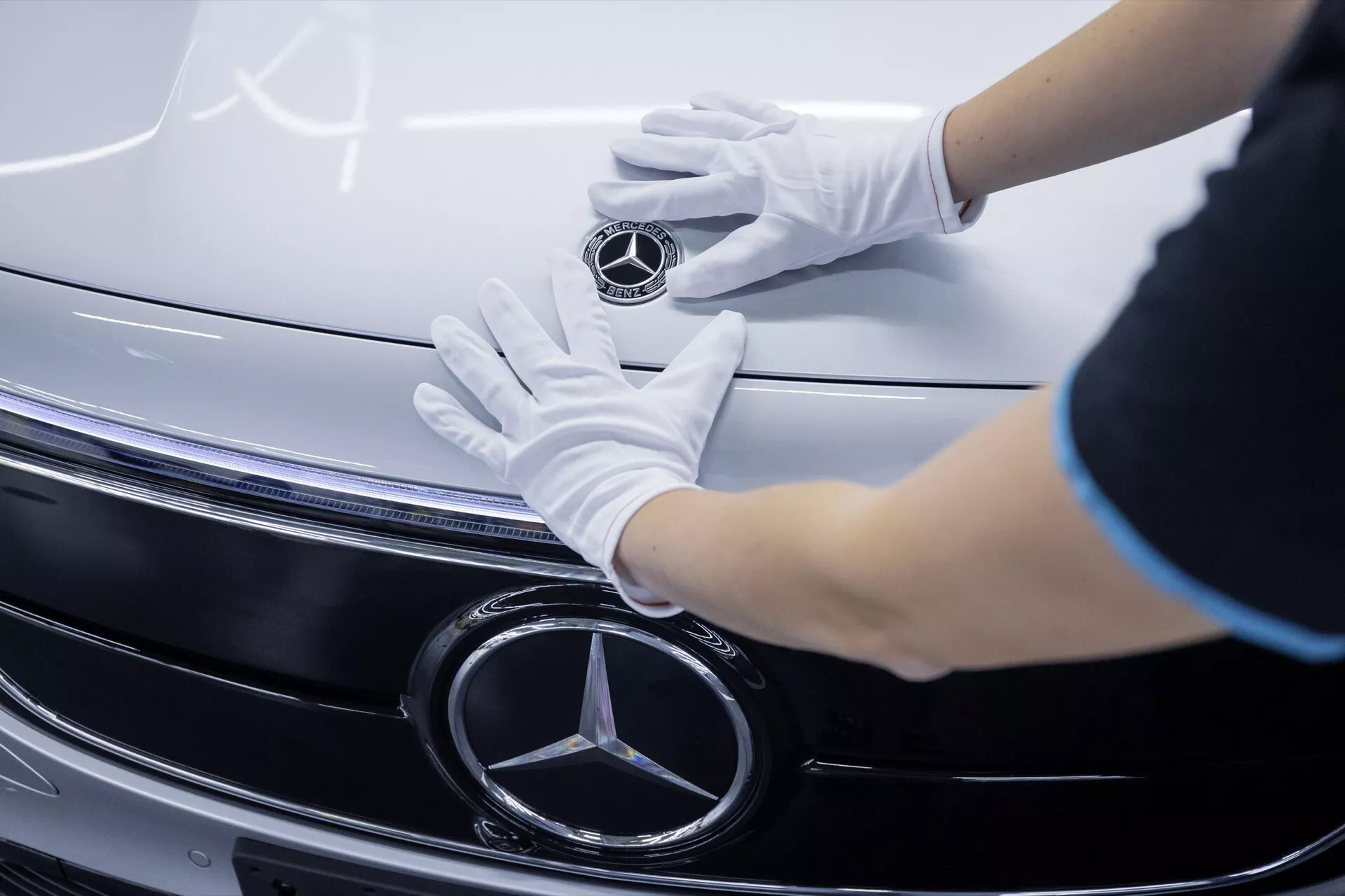 Detailní pohled na mřížku a emblém Mercedes-Benz EQA 2021 s rukama v bílých rukavicích, ukazující detaily z chromu a technologii LED světlometů.