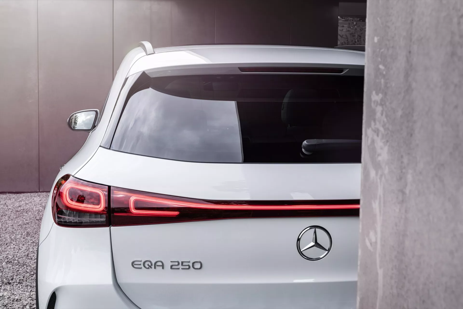 Detailní částečný pohled na bílý elektrický kompaktní SUV Mercedes-Benz EQA 250 z zadního tříčtvrtečního úhlu, prezentující prodloužené LED zadní světlo a minimalistický design s emblémem vozidla a označením EQA 250, zaparkovaný v blízkosti nevýrazné šedé plochy