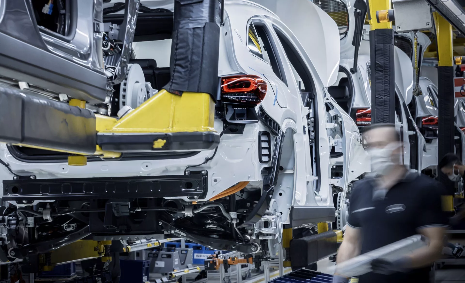 Montáž počáteční fáze Mercedes-Benz EQA 2021 na výrobní lince, zobrazující karosérii a jednolitou šasi podporované automatickými robotickými rameny s viditelnou zadní strukturou a montážními pracovníky na pozadí