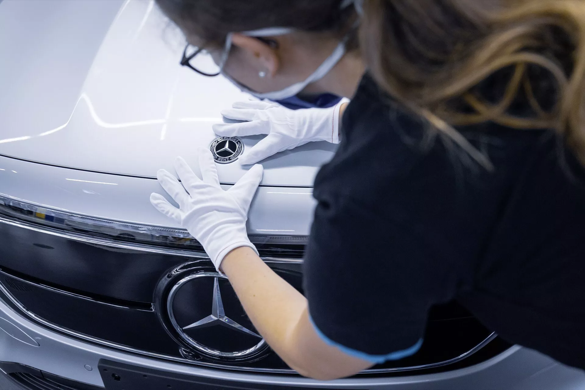 Detailní pohled na přední mřížku a emblém Mercedesu-Benz EQA 2021 s osobou v bílých rukavicích, která emblém upravuje, což zvýrazňuje prémiové zpracování a design vozidla