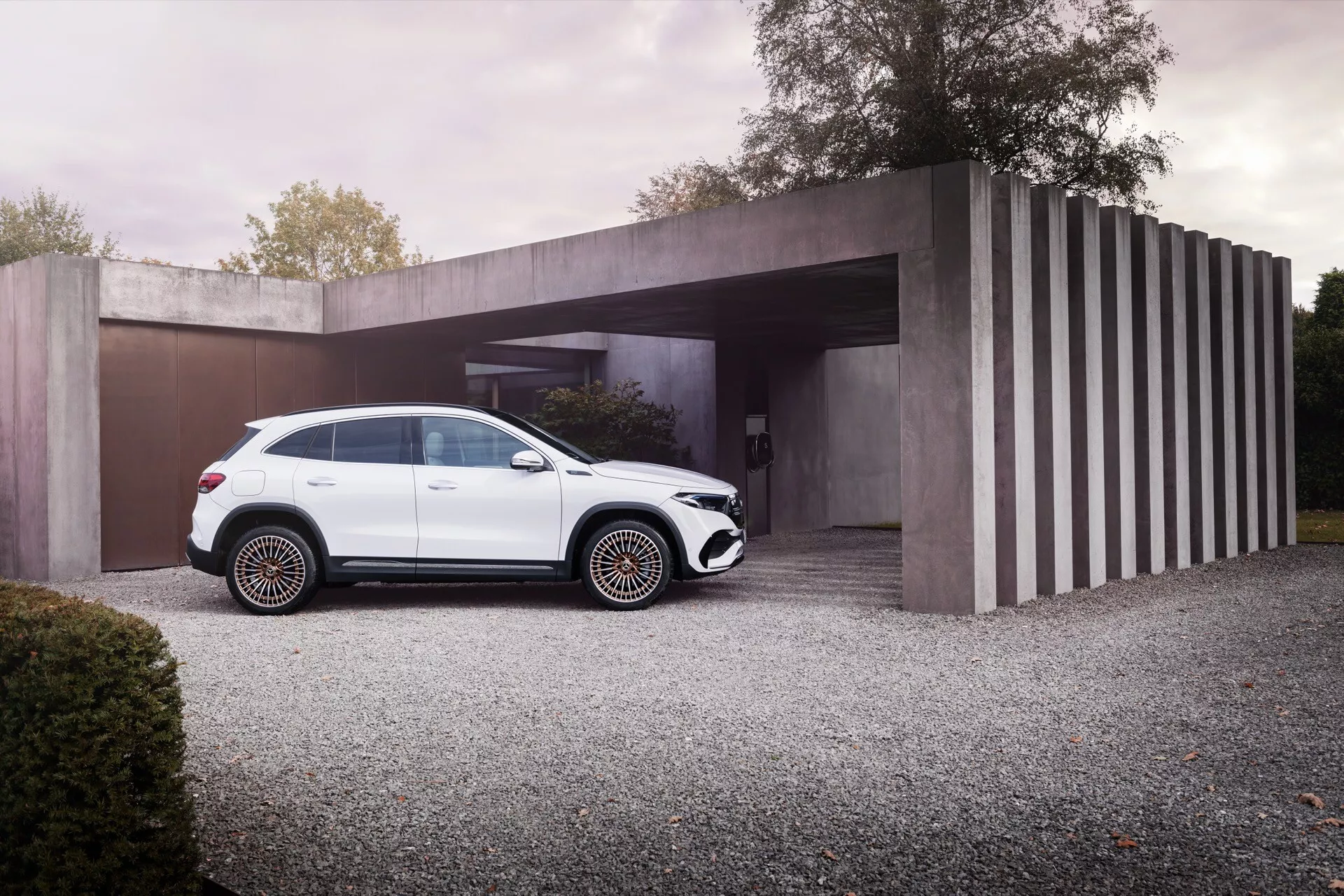 Luxusní SUV Mercedes-Benz EQA 2021 v bílé barvě zaparkované na štěrkové příjezdové cestě s moderním minimalistickým domem na pozadí, prezentující elegantní design, černé dekorativní prvky, vícepaprsková kola a střešní nosiče