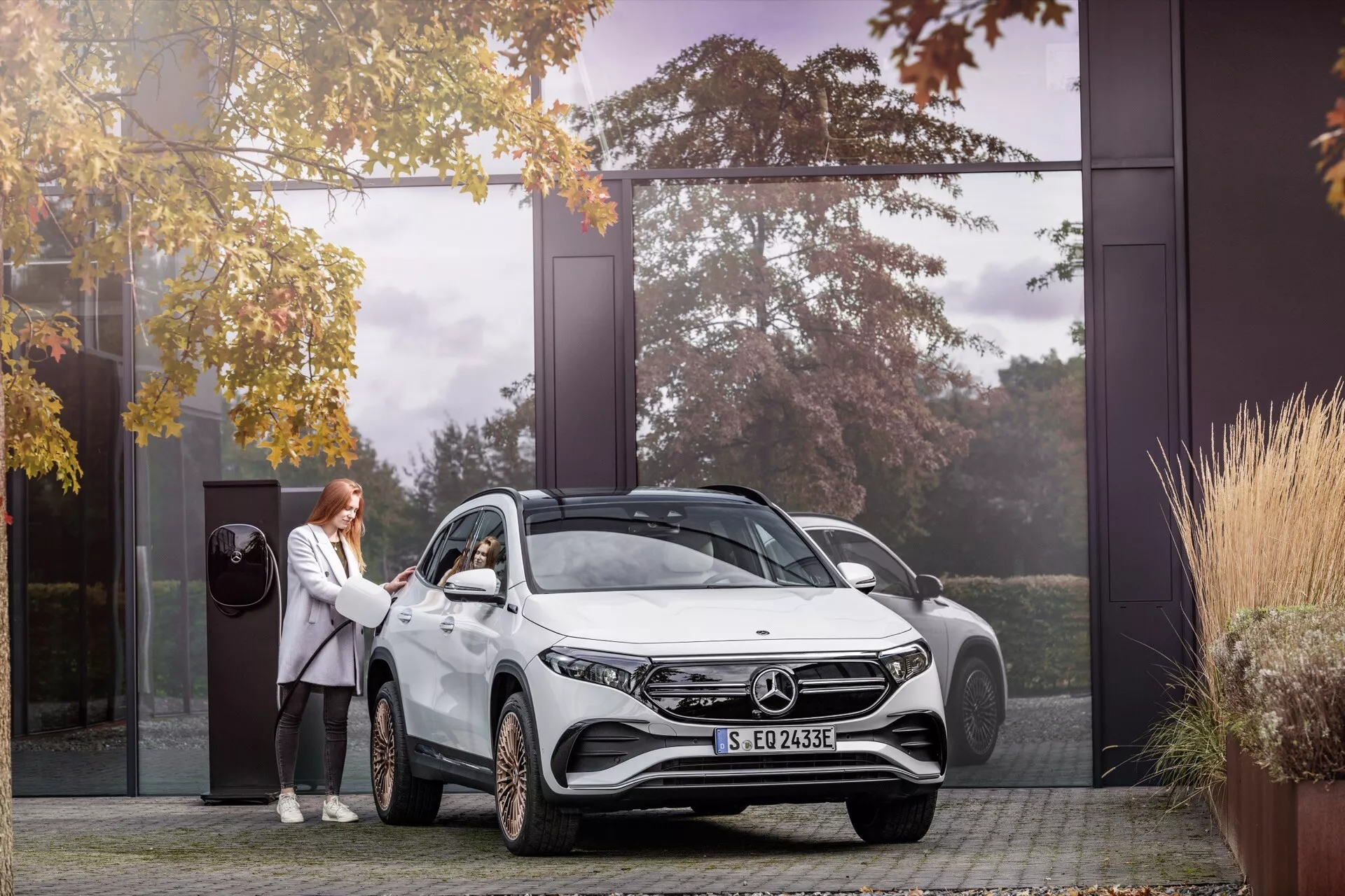 SUV Mercedes-Benz EQA 2021 v příjezdové cestě obytného domu, s LED světlomety, slitinovými koly, elektrickým označením a osobou demonstrující bezklíčový vstup, obklopeno podzimními stromy