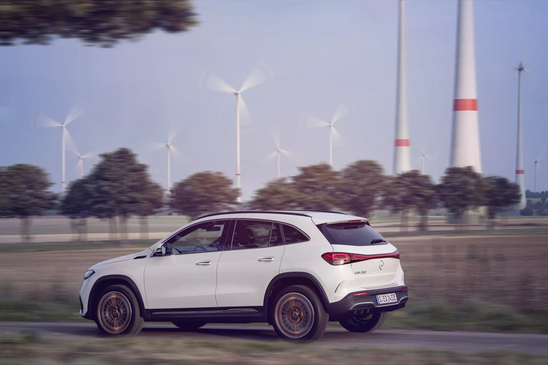 Dynamický pohyb Mercedes-Benz EQA 2021, profilový boční pohled na moderní SUV na zpevněné silnici s větrnými turbínami na pozadí, předvádějící aerodynamický design, LED zadní světla a vysokou světlou výšku