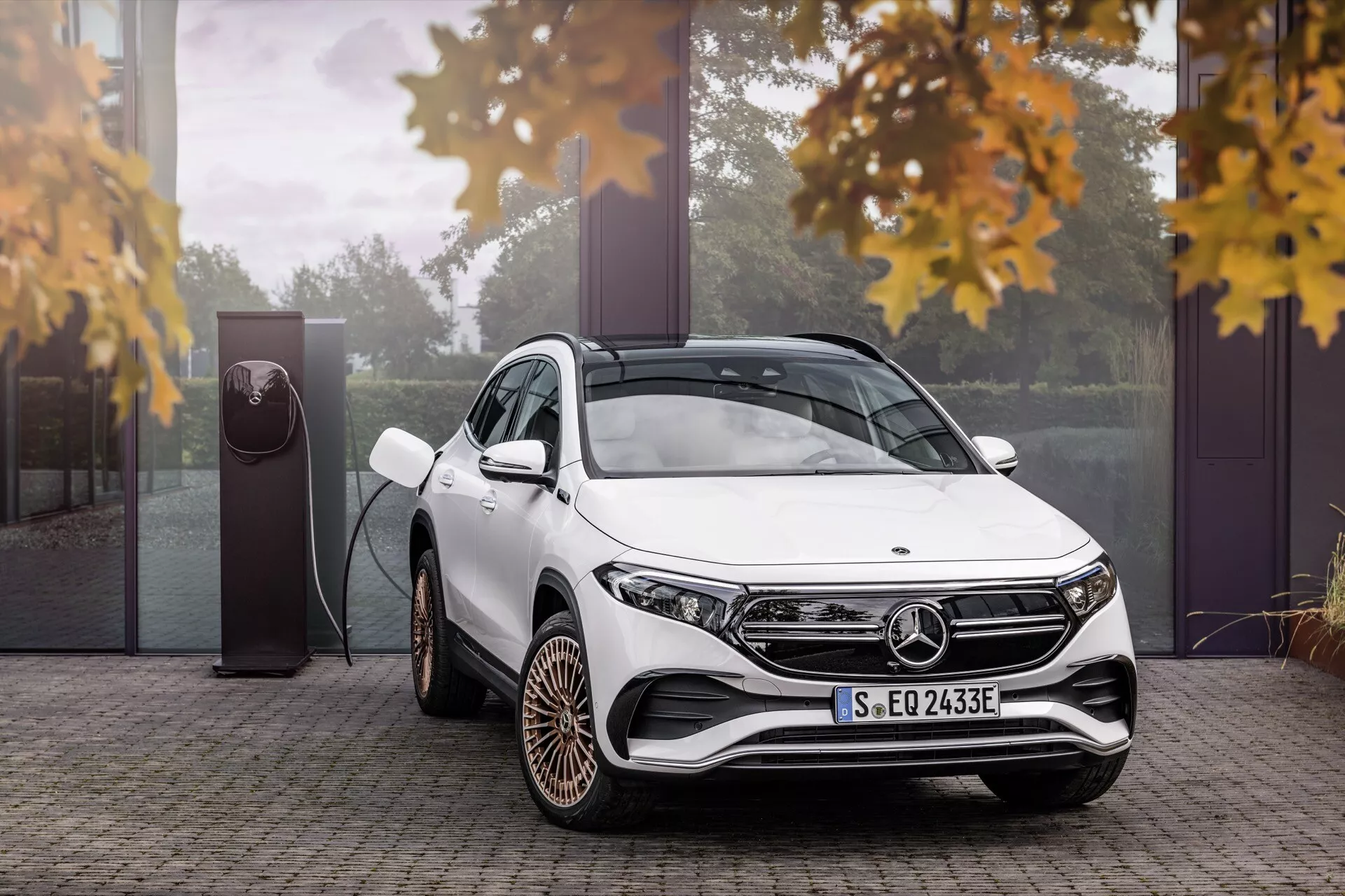 Bílé luxusní SUV Mercedes-Benz EQA 243 roku 2021 se zobrazením přední a boční části v trojčtvrtě pohledu, zdůrazňující moderní LED světlomety, charakteristická slitinová kola a technologii nabíjení elektrického vozu, obklopená náznakem podzimního listí, což zdůrazňuje jeho ekologický design v luxusním venkovním prostředí