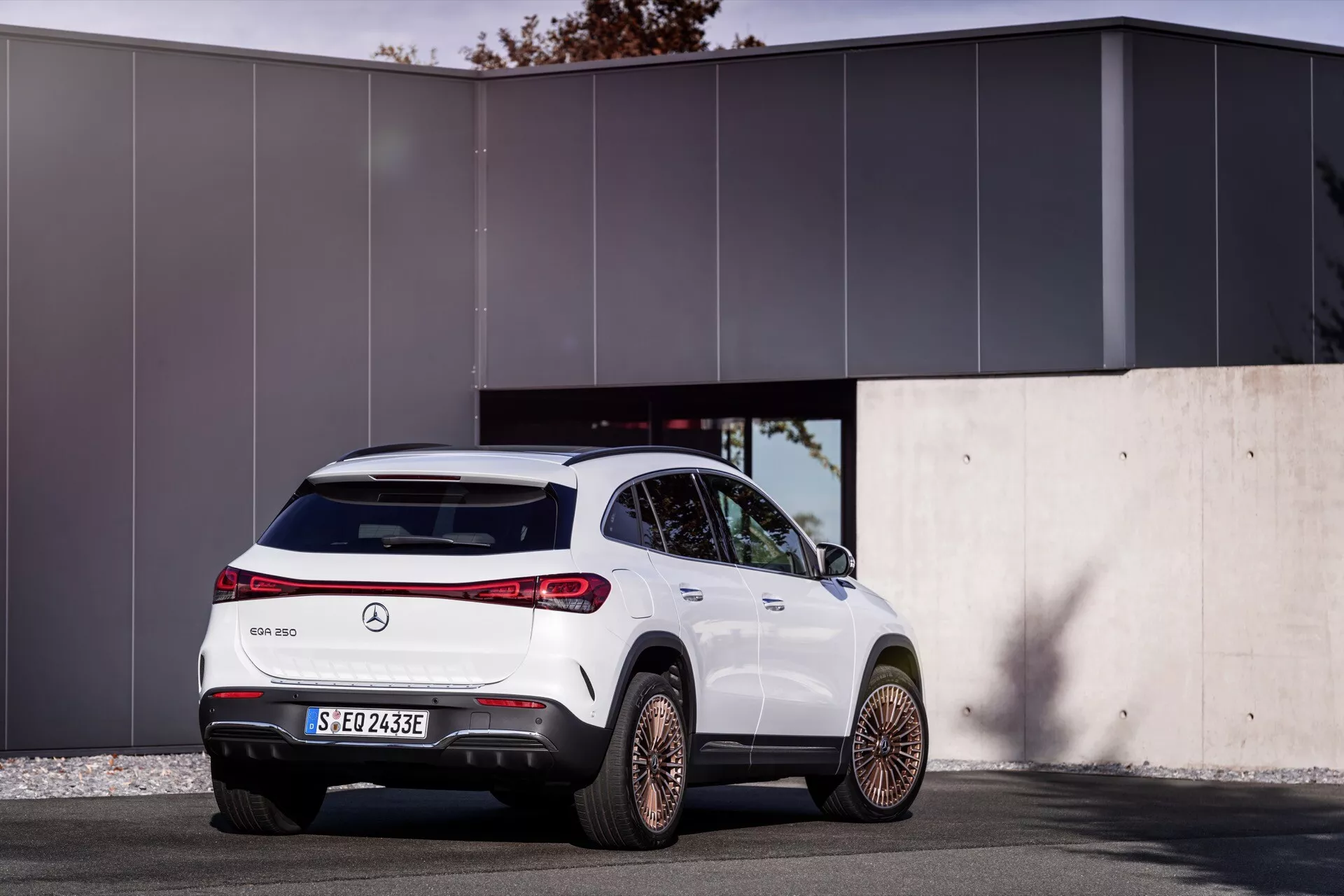 Tříčtvrteční zadní pohled na bílé SUV Mercedes-Benz EQA 2021 zaparkované venku, s moderním designem s konturovanými liniemi, charakteristickými obklopujícími zadními světly, sportovním zadním nárazníkem s difuzorem, slitinovými koly se pěti rozdělenými paprsky, označením elektrického vozidla, parkovacími senzory vzadu, vysoko umístěnou stopovou lampou, lesklým povrchem s chromovými lištami, na pozadí minimalistické moderní budovy na slunci.