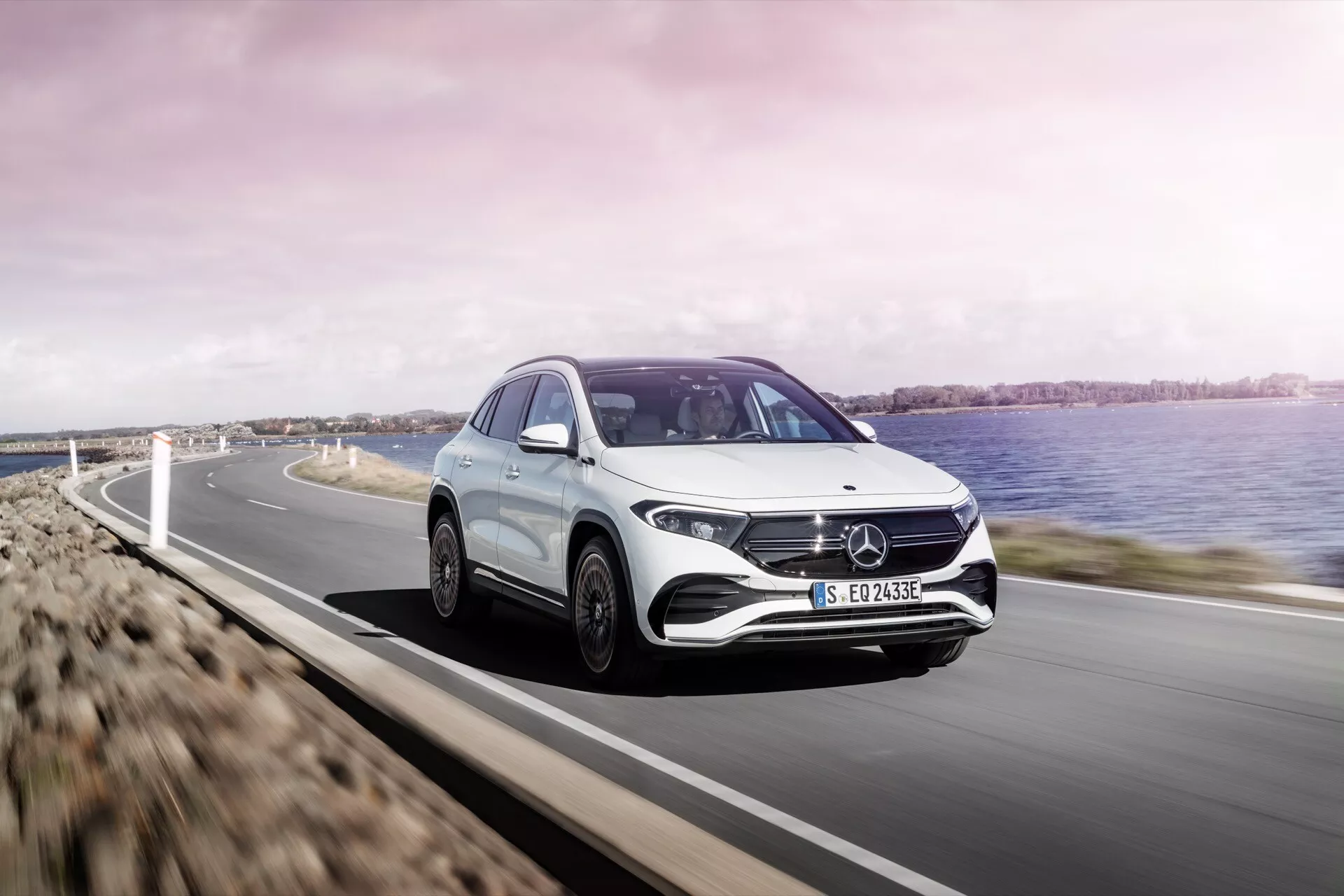 Mercedes-Benz EQA 2021 v pohybu na pobřežní silnici, předvádějící aerodynamický design, úhlová LED světla a charakteristickou masku s emblémem, viditelný proti malebnému pozadí s vodou
